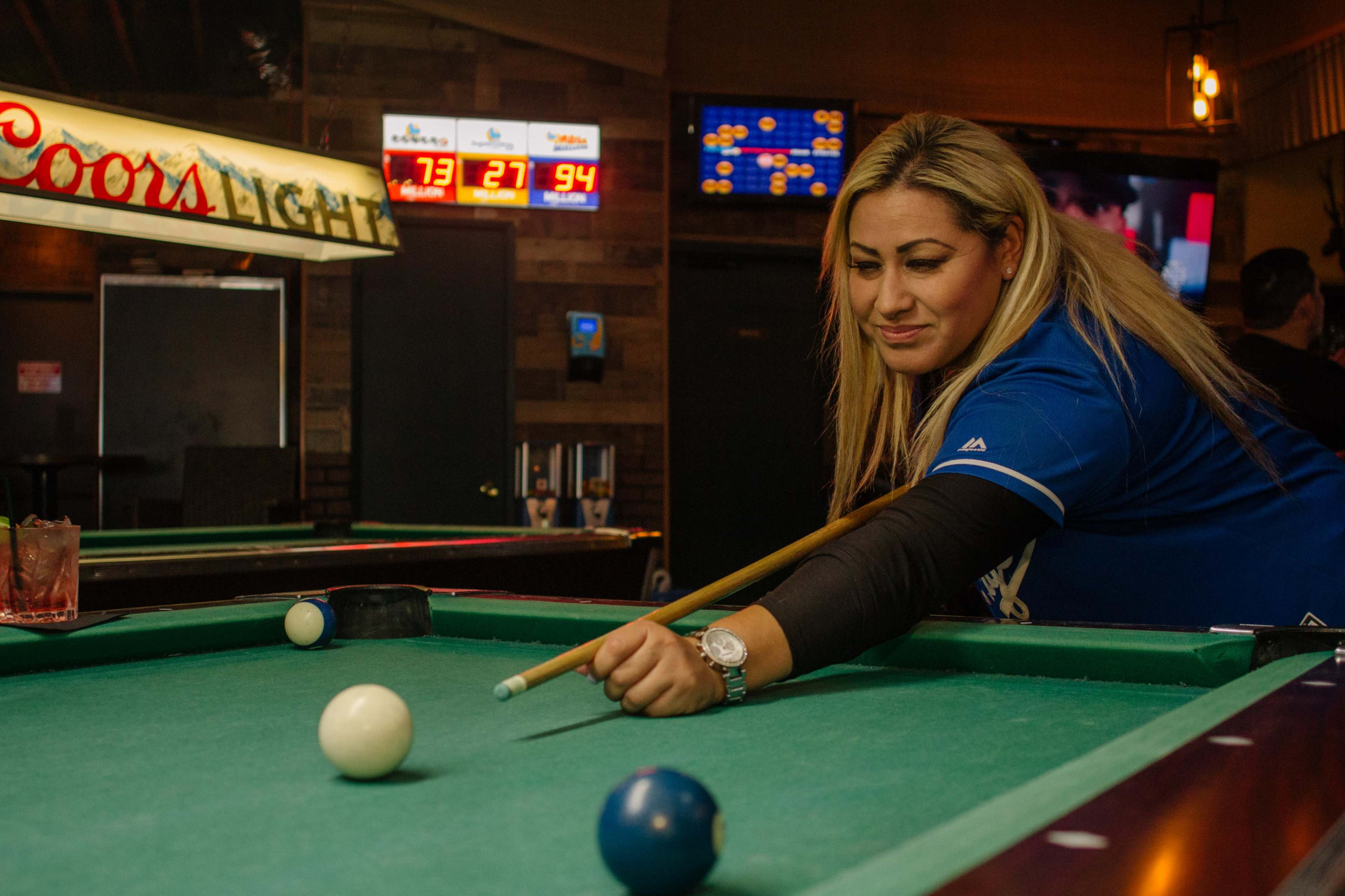 A woman in a blue shirt is leaning over a pool table, aiming with a cue stick at a white ball while a scoreboard and neon signs are visible in the background.