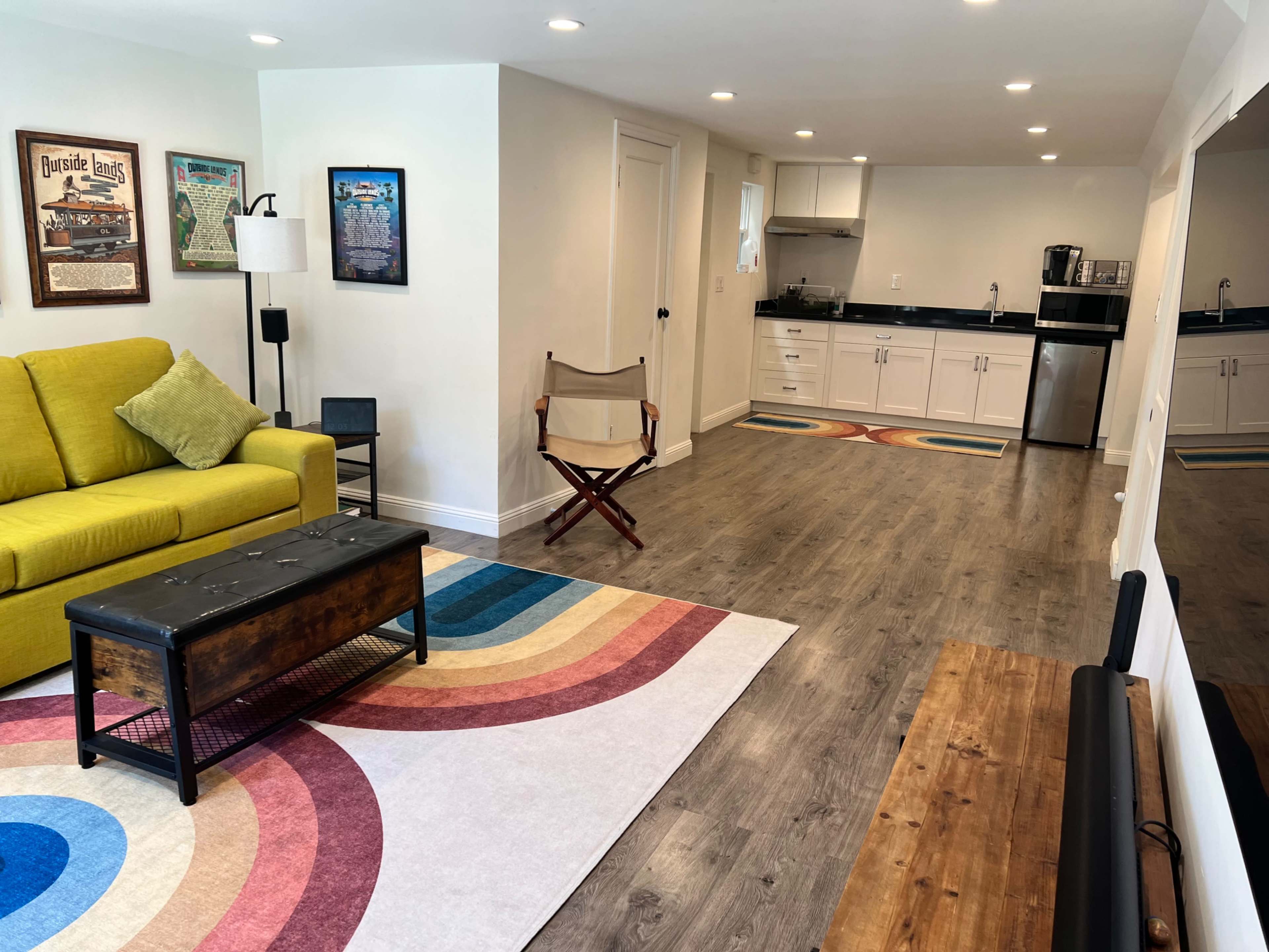 A modern living space featuring a yellow couch, a coffee table, a chair, and a colorful area rug, with a kitchenette in the background.