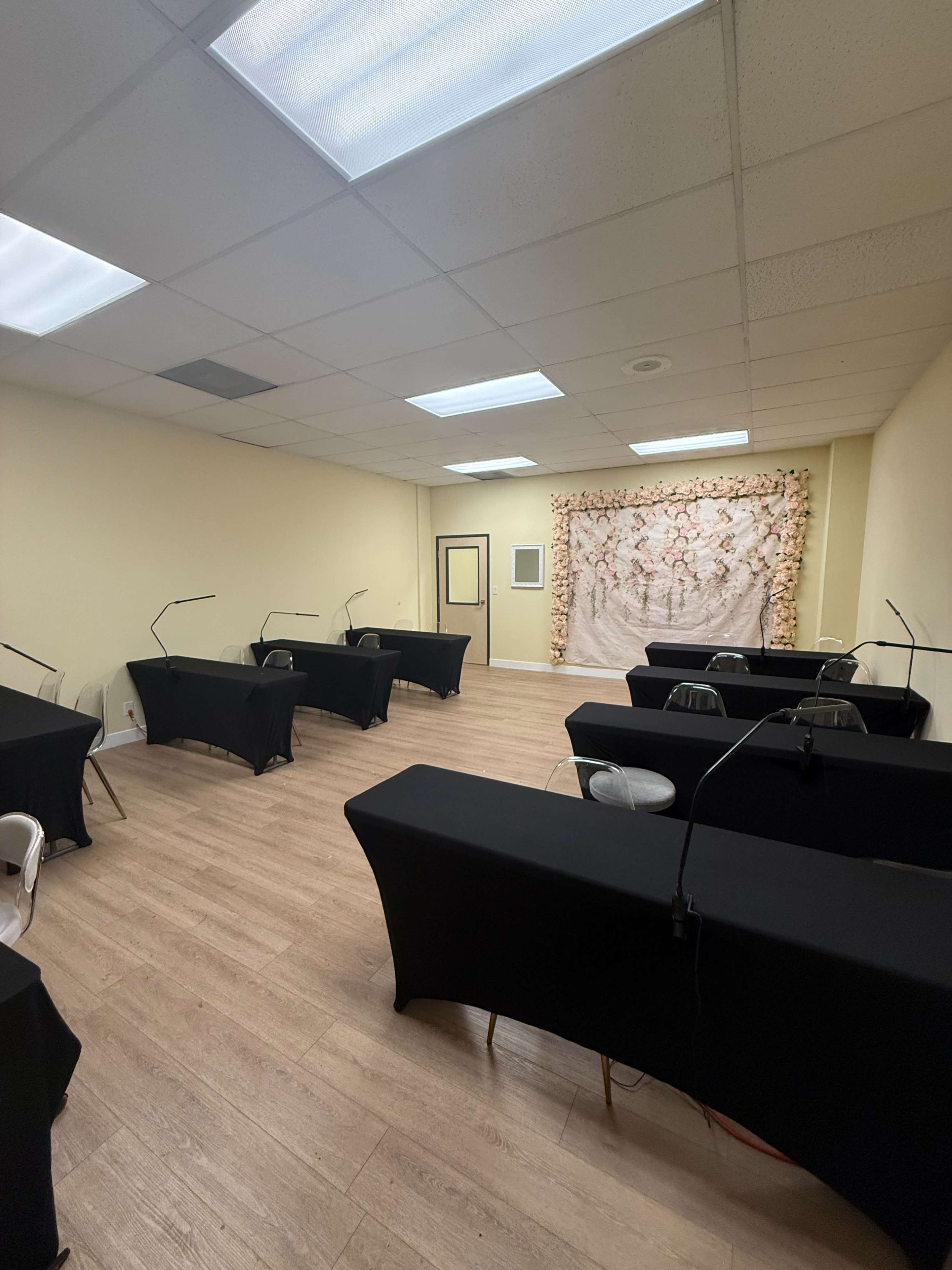 The image shows a spacious room with several black tables arranged in a grid pattern, each equipped with lamps, and a floral backdrop on one wall.