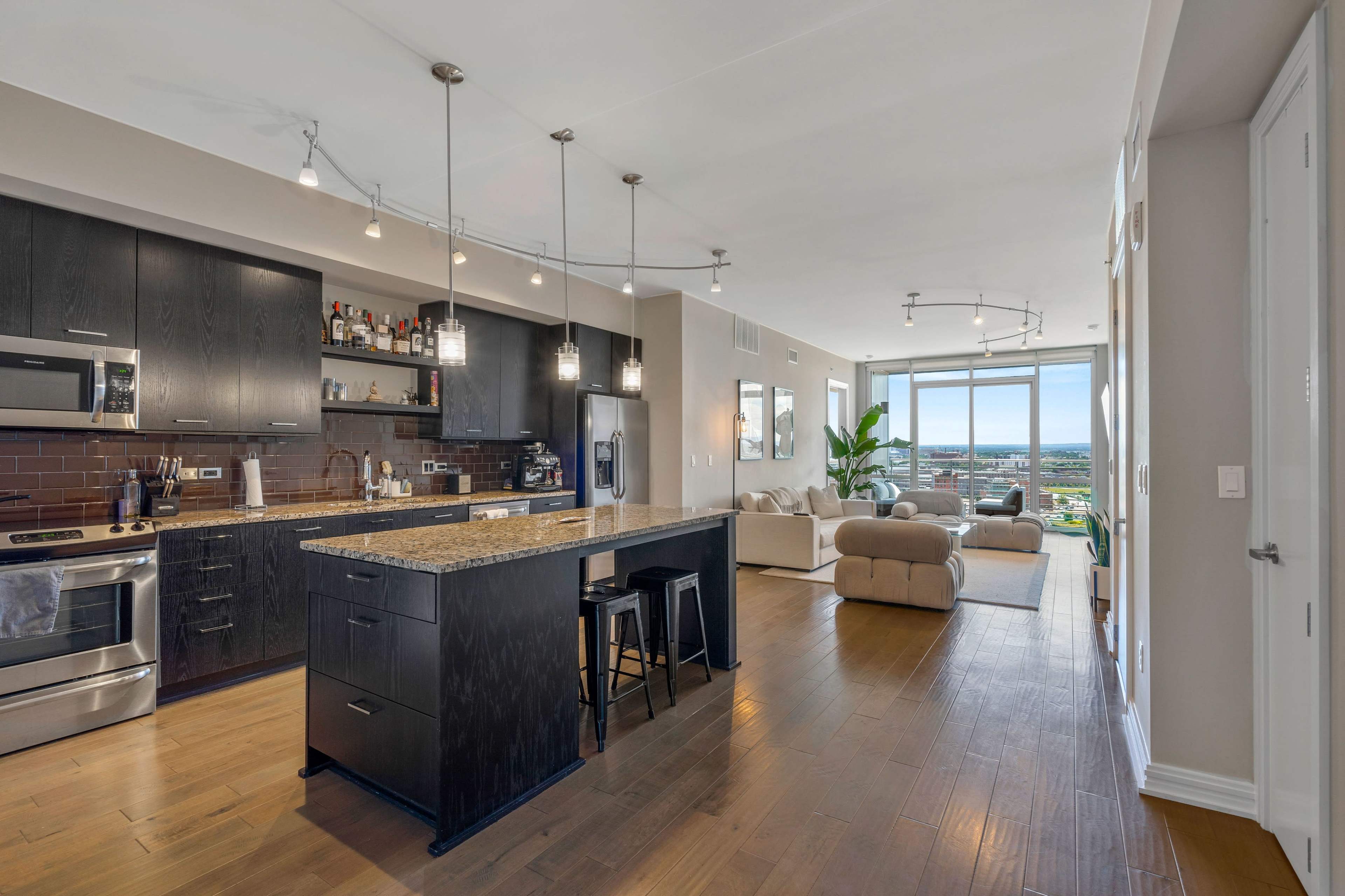 A modern kitchen with dark cabinetry and a granite countertop, leading to a bright living area with a large window and a view of the skyline.