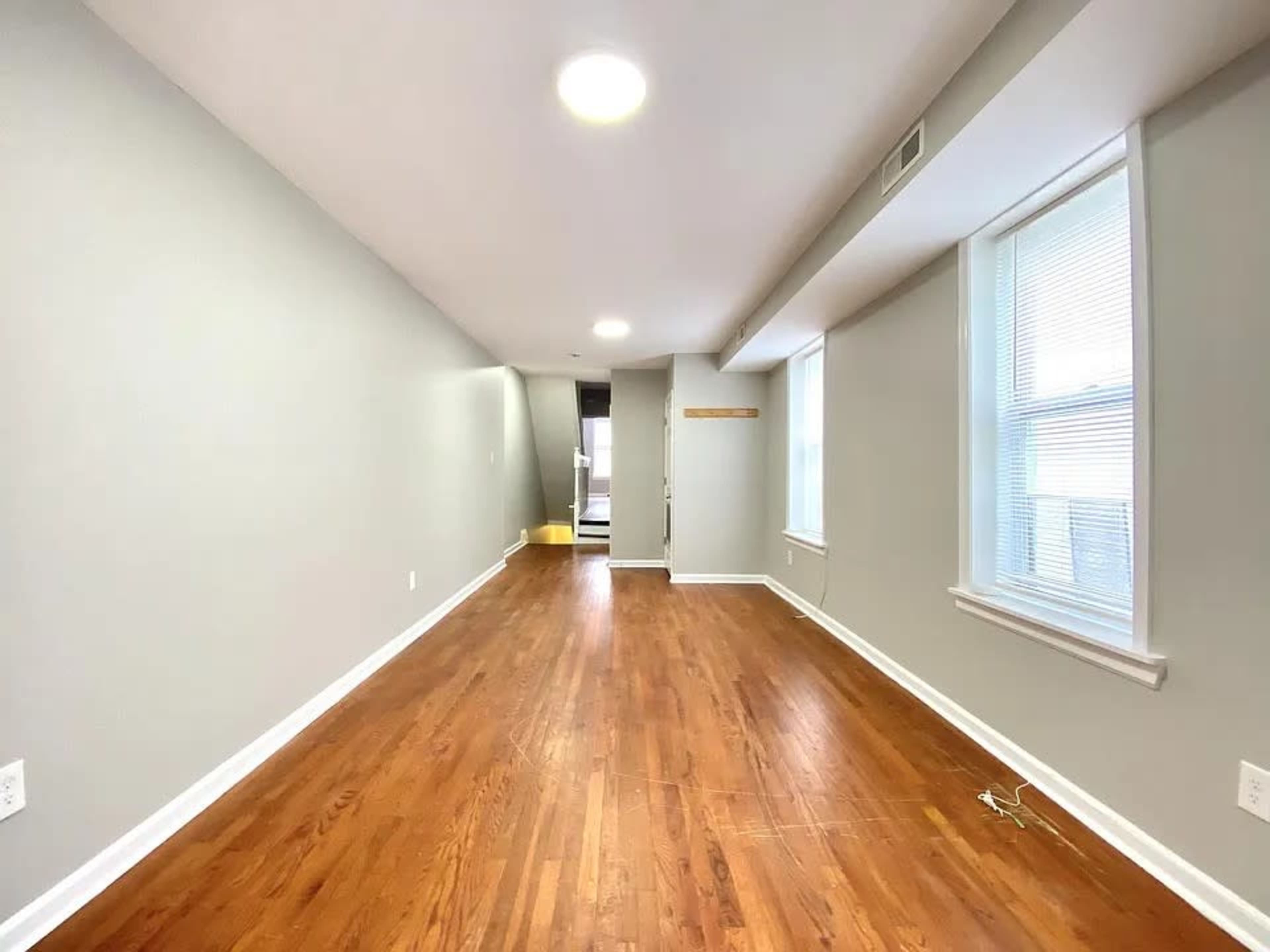 The image shows a vacant room with polished hardwood floors, gray walls, and two windows providing natural light.