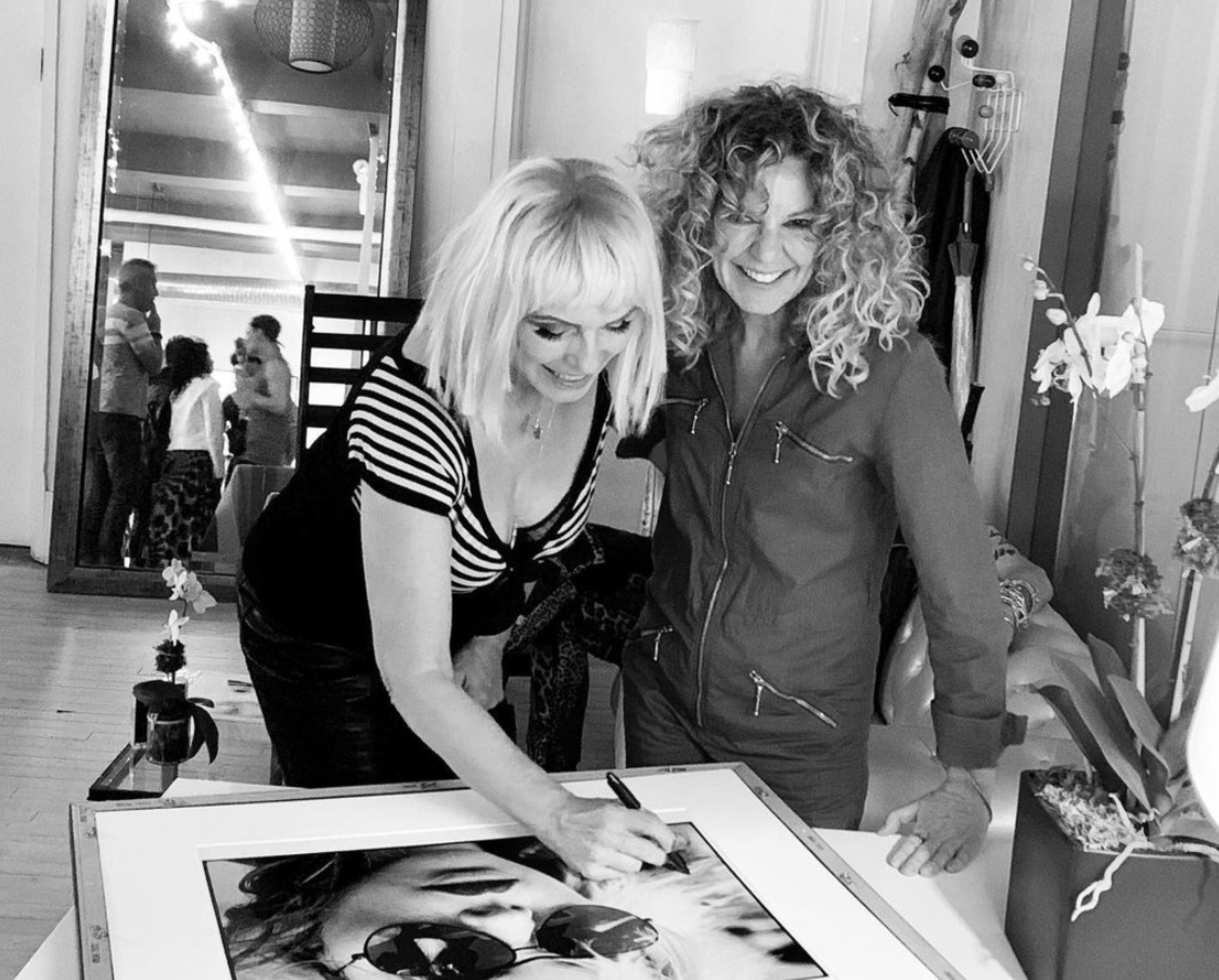 Two women are engaged in signing a framed black-and-white photograph in a well-lit interior space.