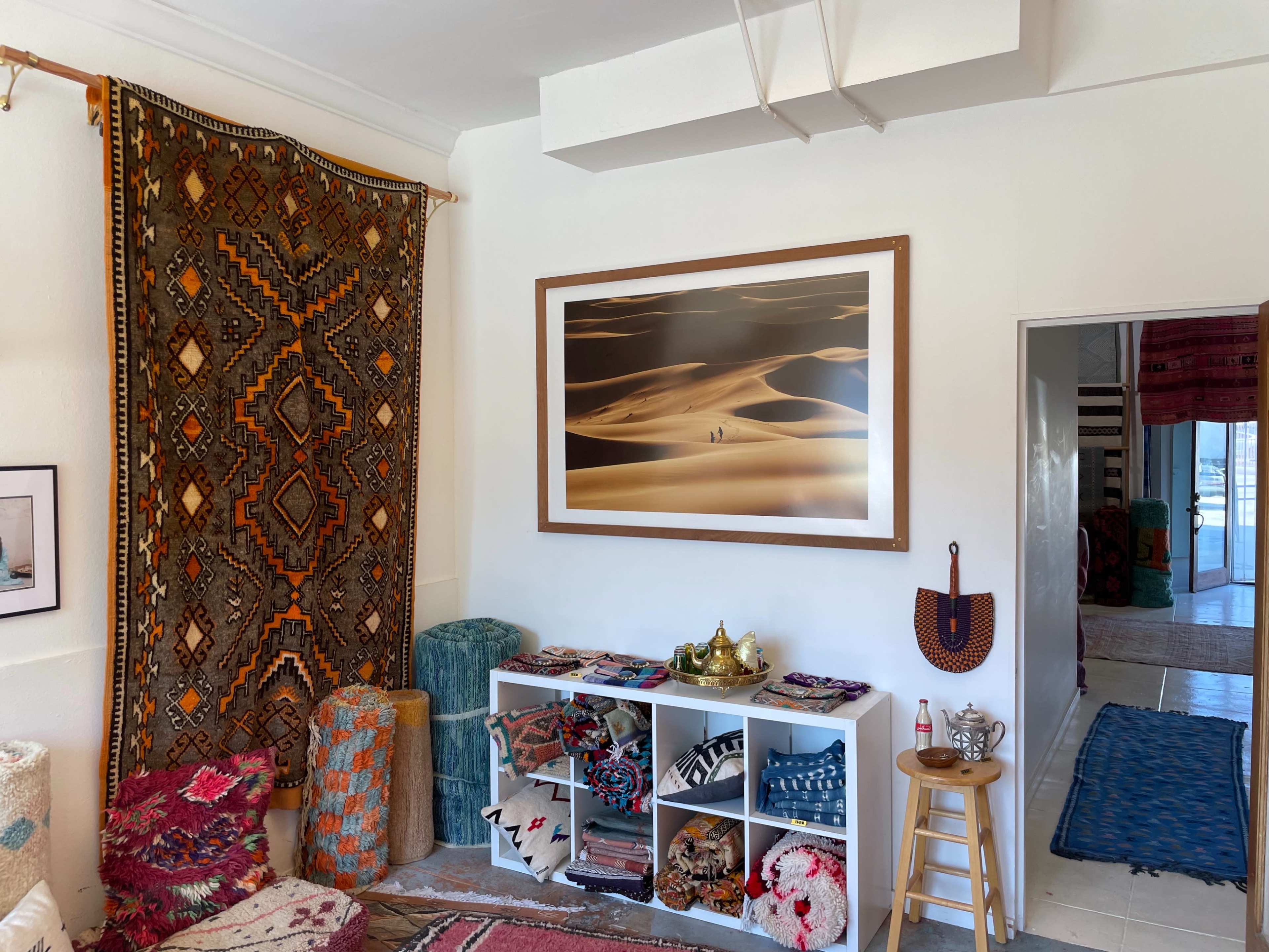 The image showcases a brightly decorated living space with a large, patterned wall hanging, a framed landscape photograph, and shelves filled with various textiles and decorative items.