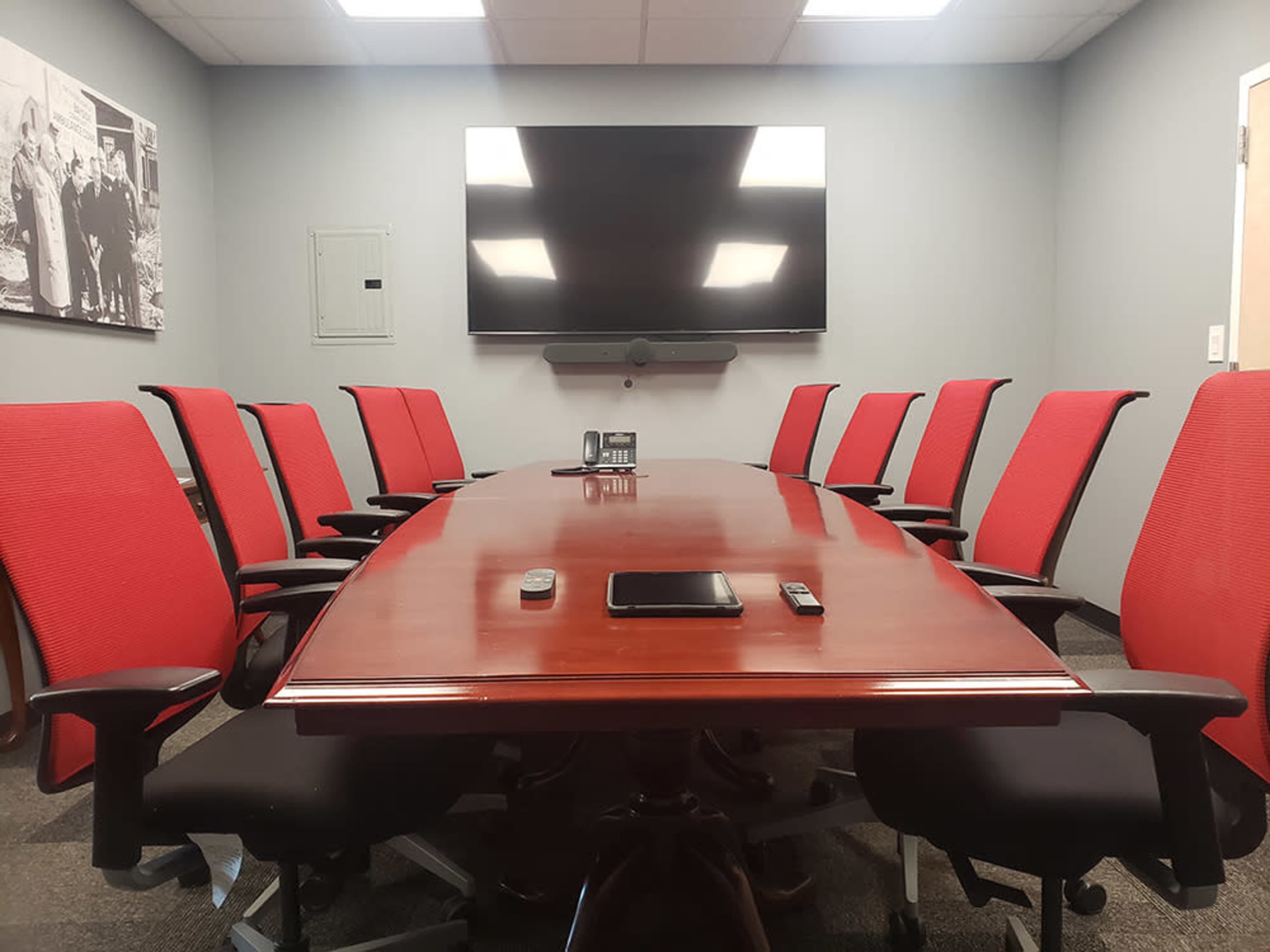 A conference room features a long wooden table surrounded by ten red chairs and a large wall-mounted TV.