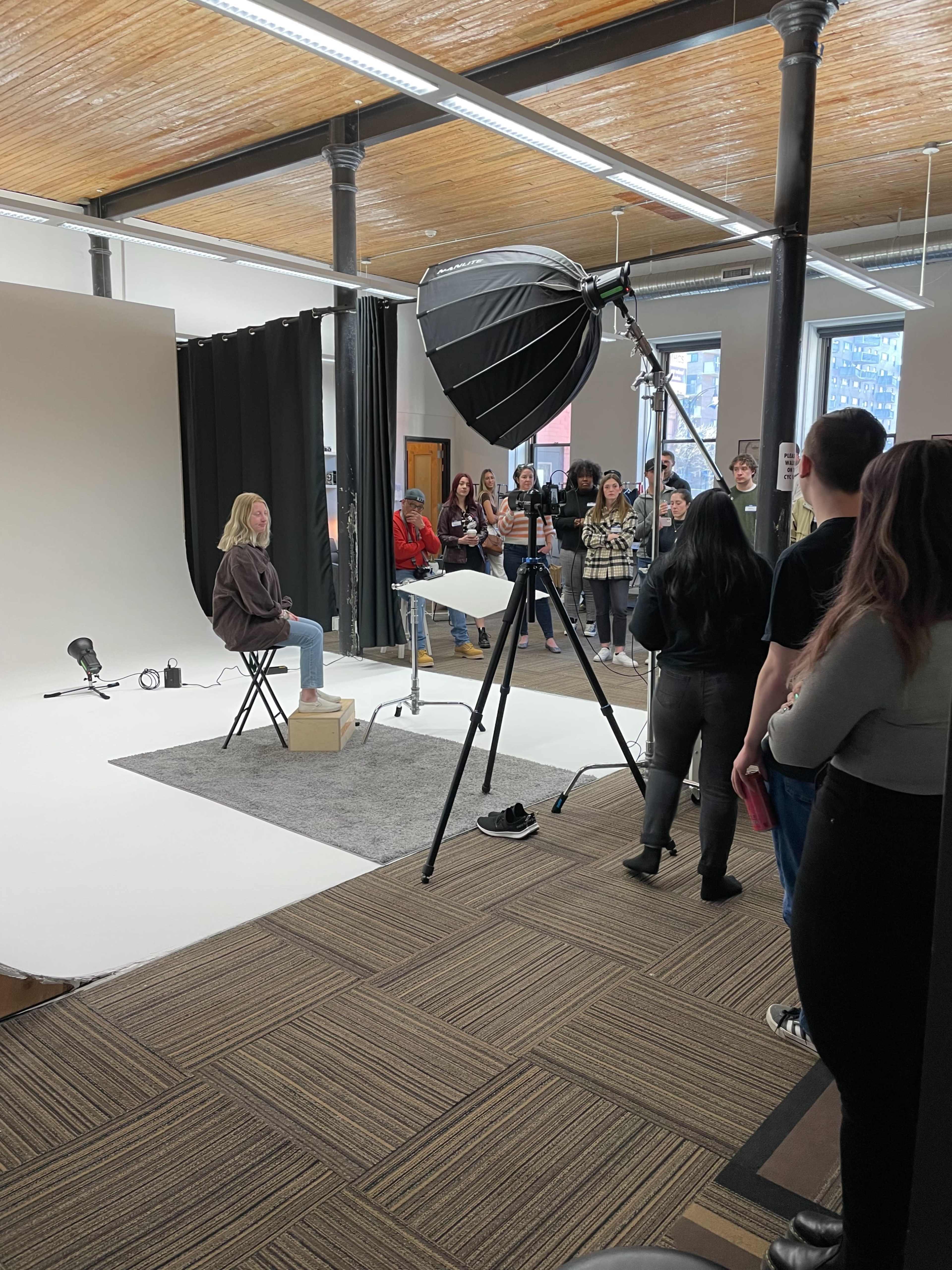 A group of people observes a photoshoot setup featuring a seated model in front of a lighting umbrella against a neutral backdrop.