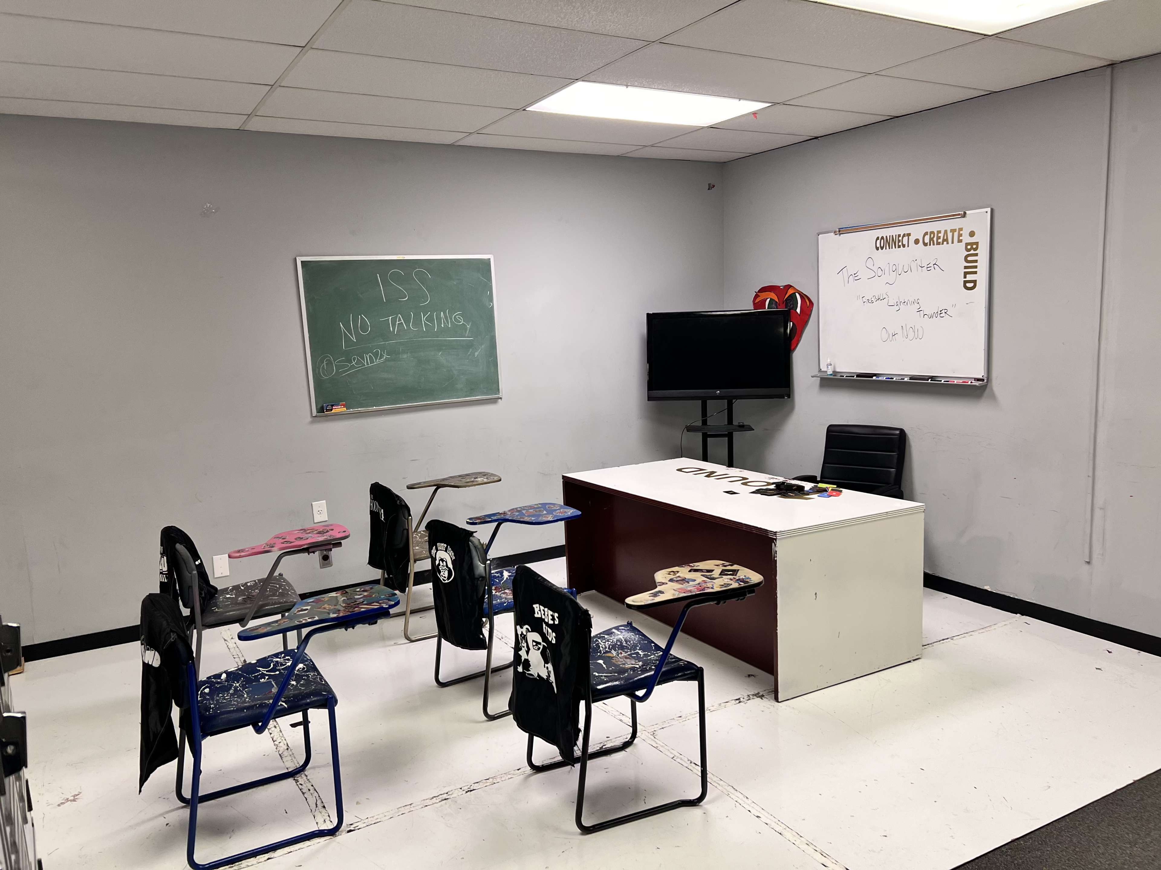 A sparse classroom with a chalkboard, a whiteboard, a desk, a chair, and several empty chairs arranged around the room.