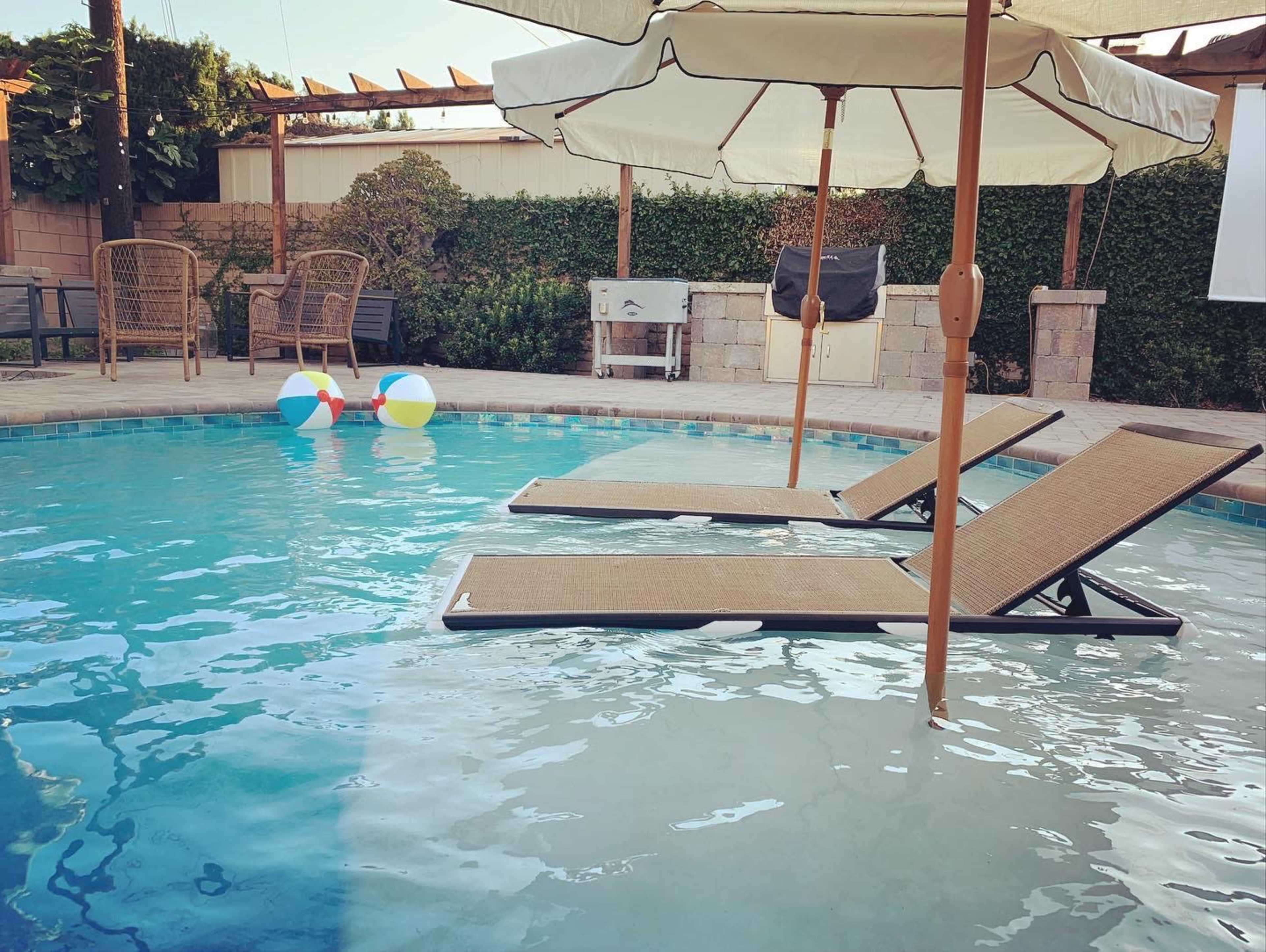 The scene shows a swimming pool with two lounge chairs partially submerged, colorful beach balls floating nearby, and an umbrella providing shade.