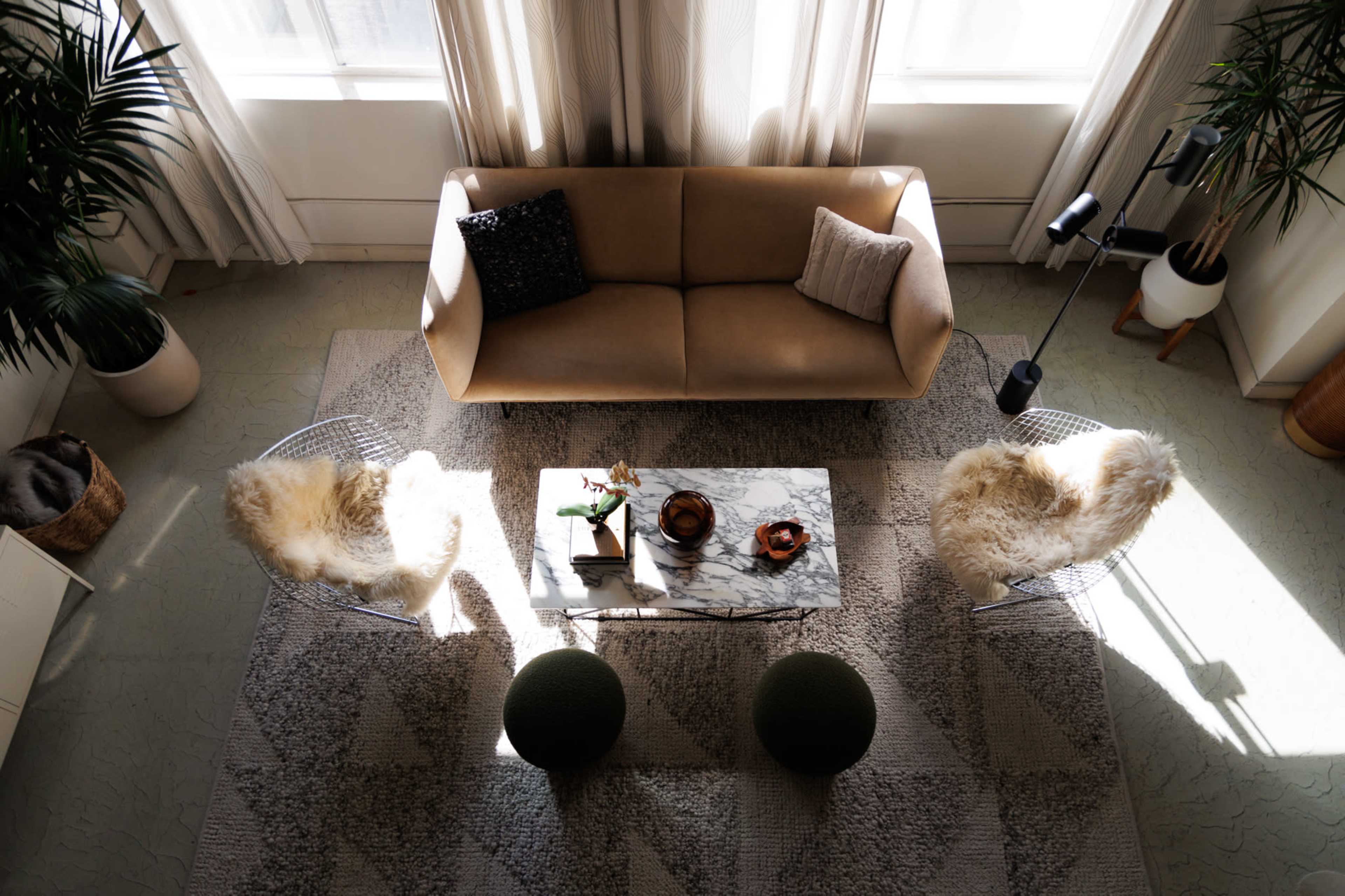 The image shows a living room with a beige sofa, two chair with fur throws, a marble coffee table, and green poufs, surrounded by plants and large windows.