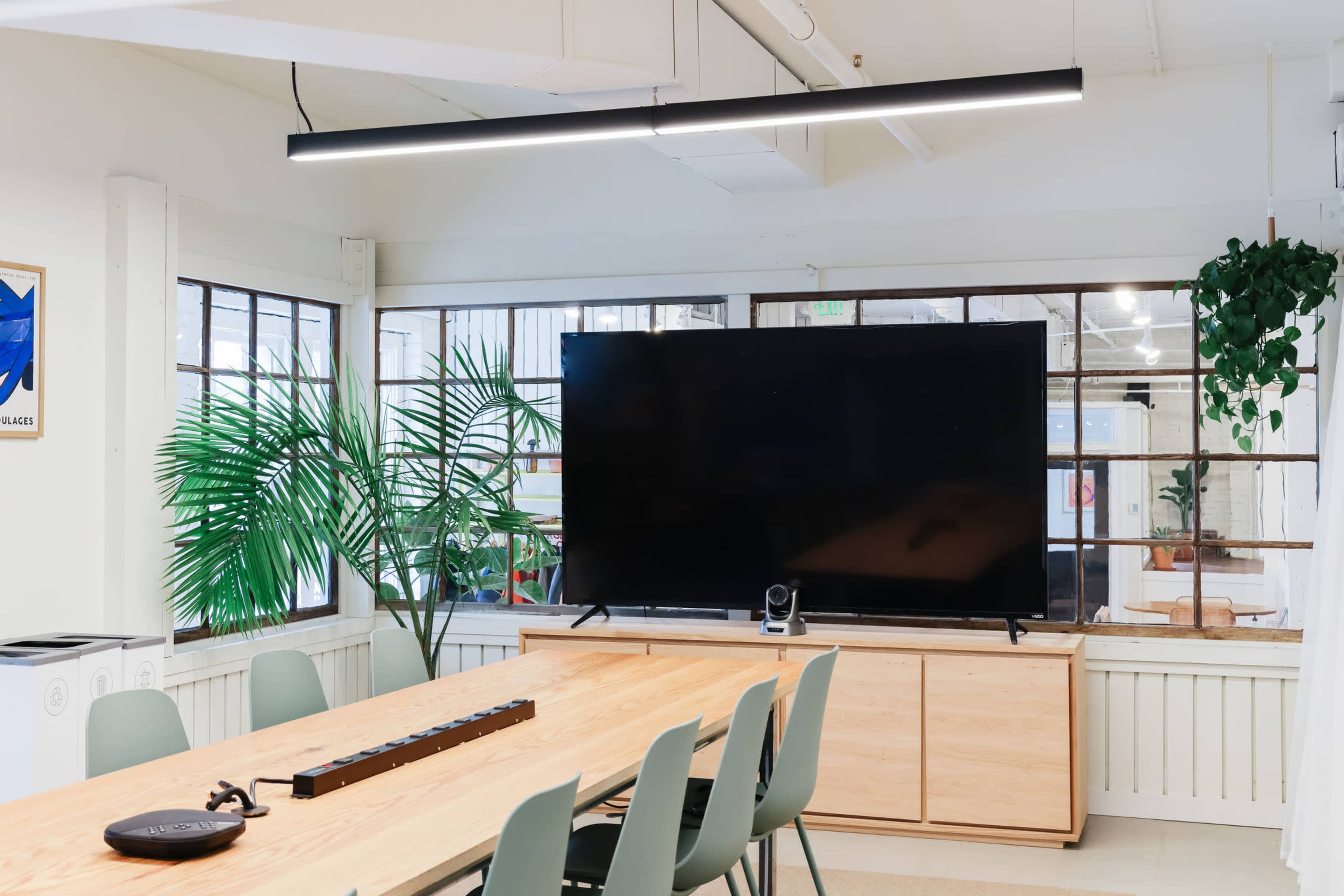 A modern conference room features a long wooden table, green chairs, a large television mounted on a cabinet, and large windows with plants.