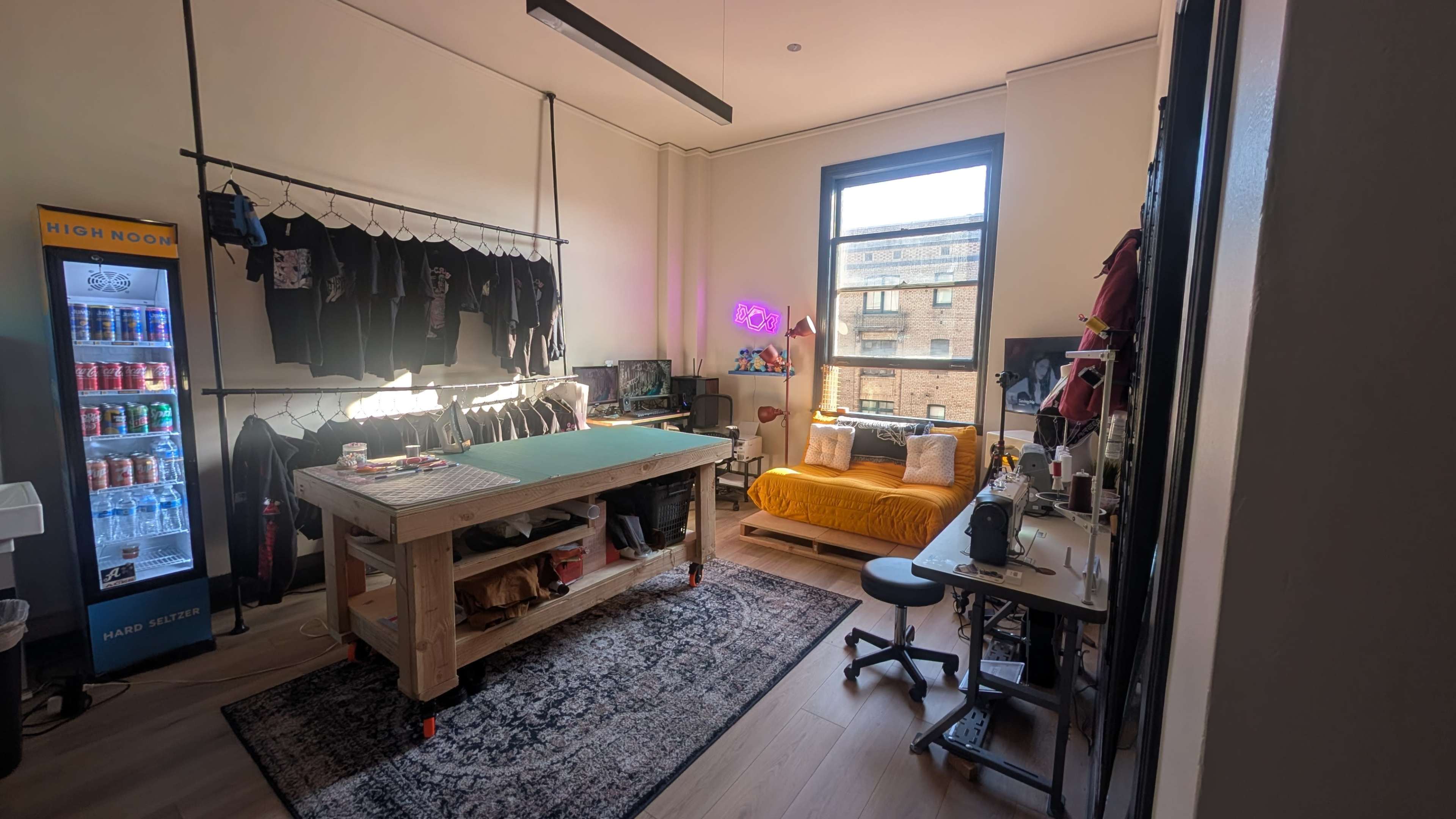 The image shows a workspace featuring a large wooden table in the center, a yellow couch, shelves with black clothing, a neon sign, and a vending machine against the wall.