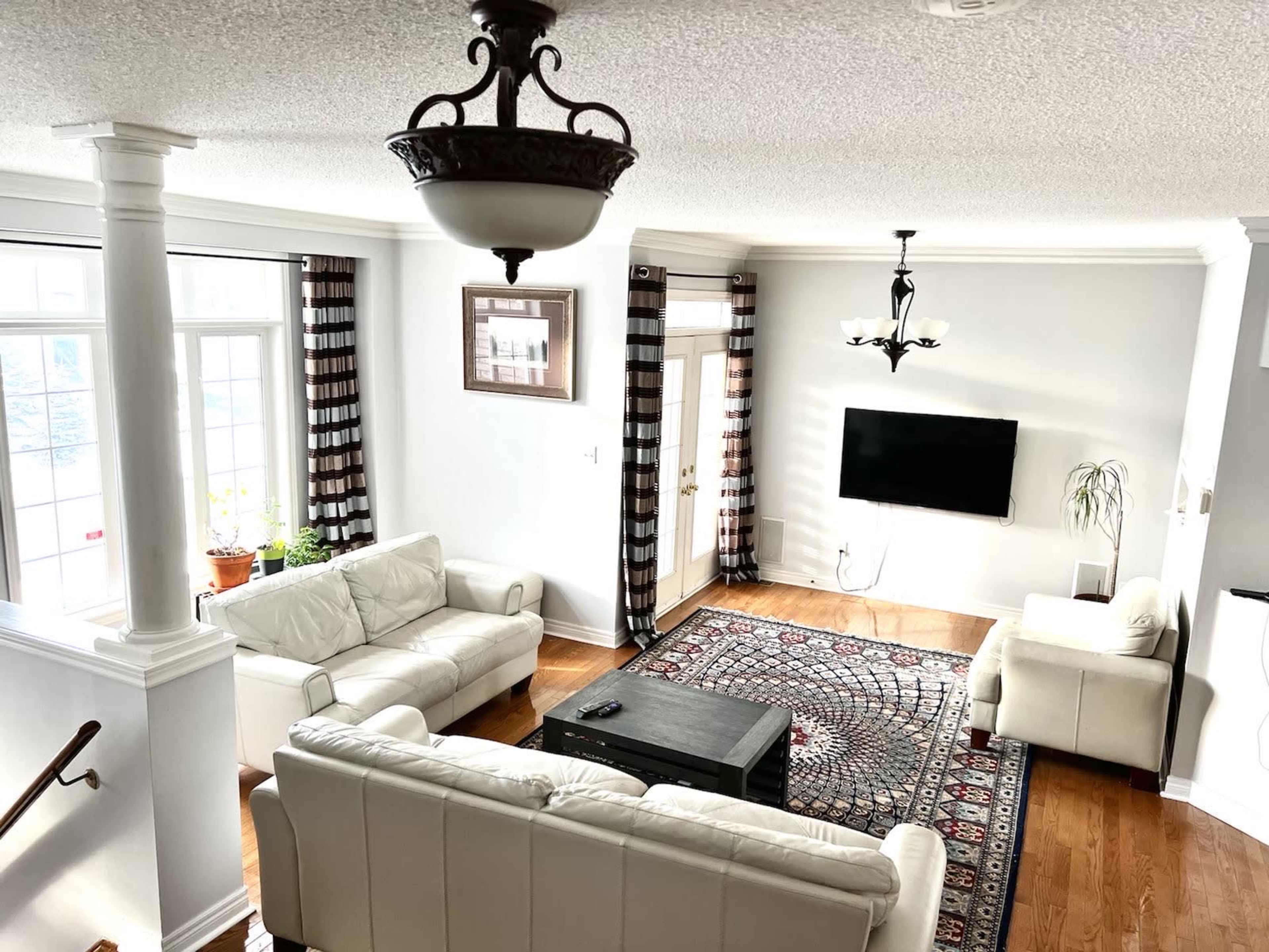 A spacious living room features a pair of white sofas, a dark coffee table, a patterned rug, and a wall-mounted television under a chandelier.