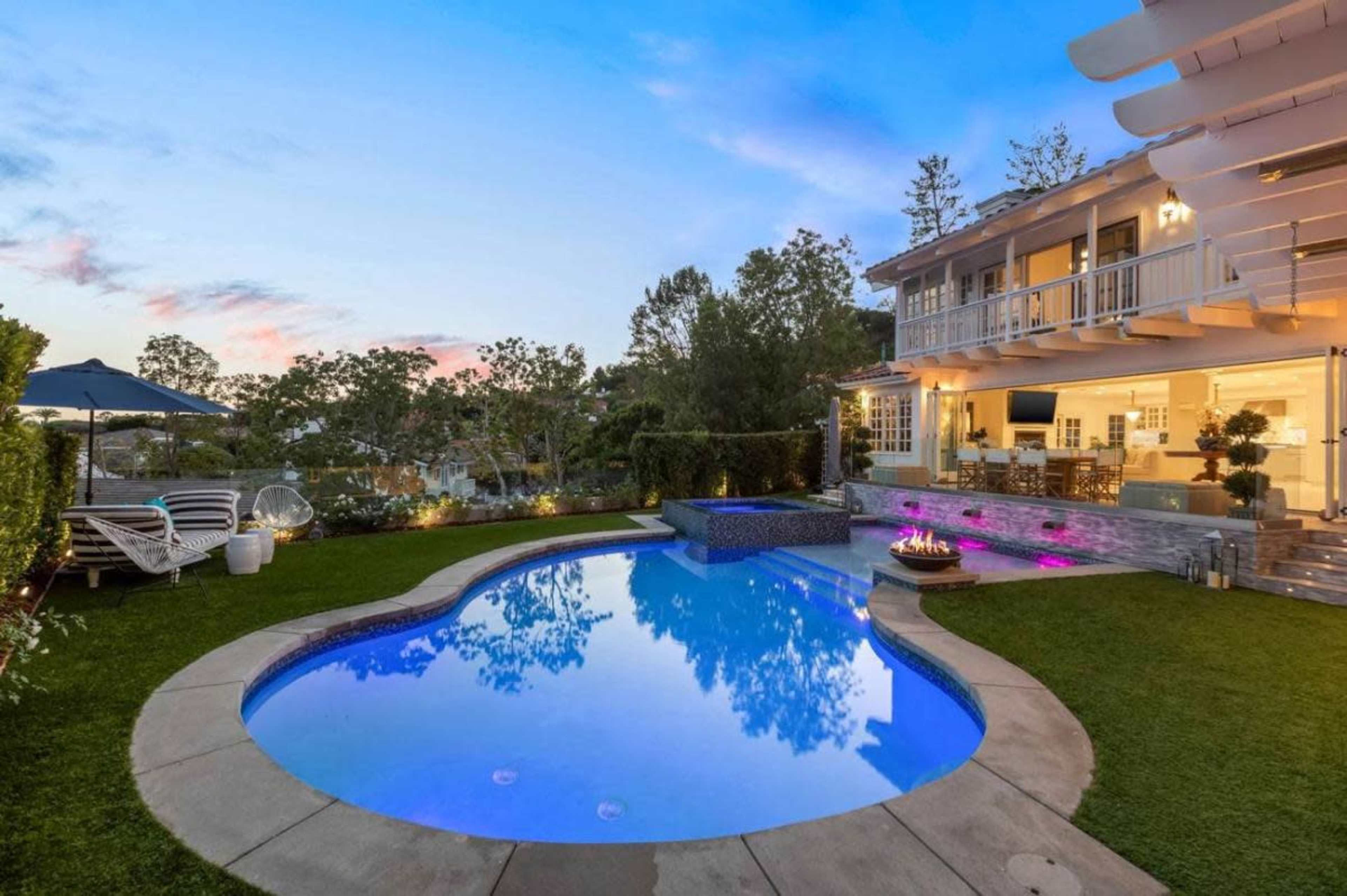 A landscaped backyard features a pool with a spa, surrounded by greenery and patio furniture, alongside a two-story house.