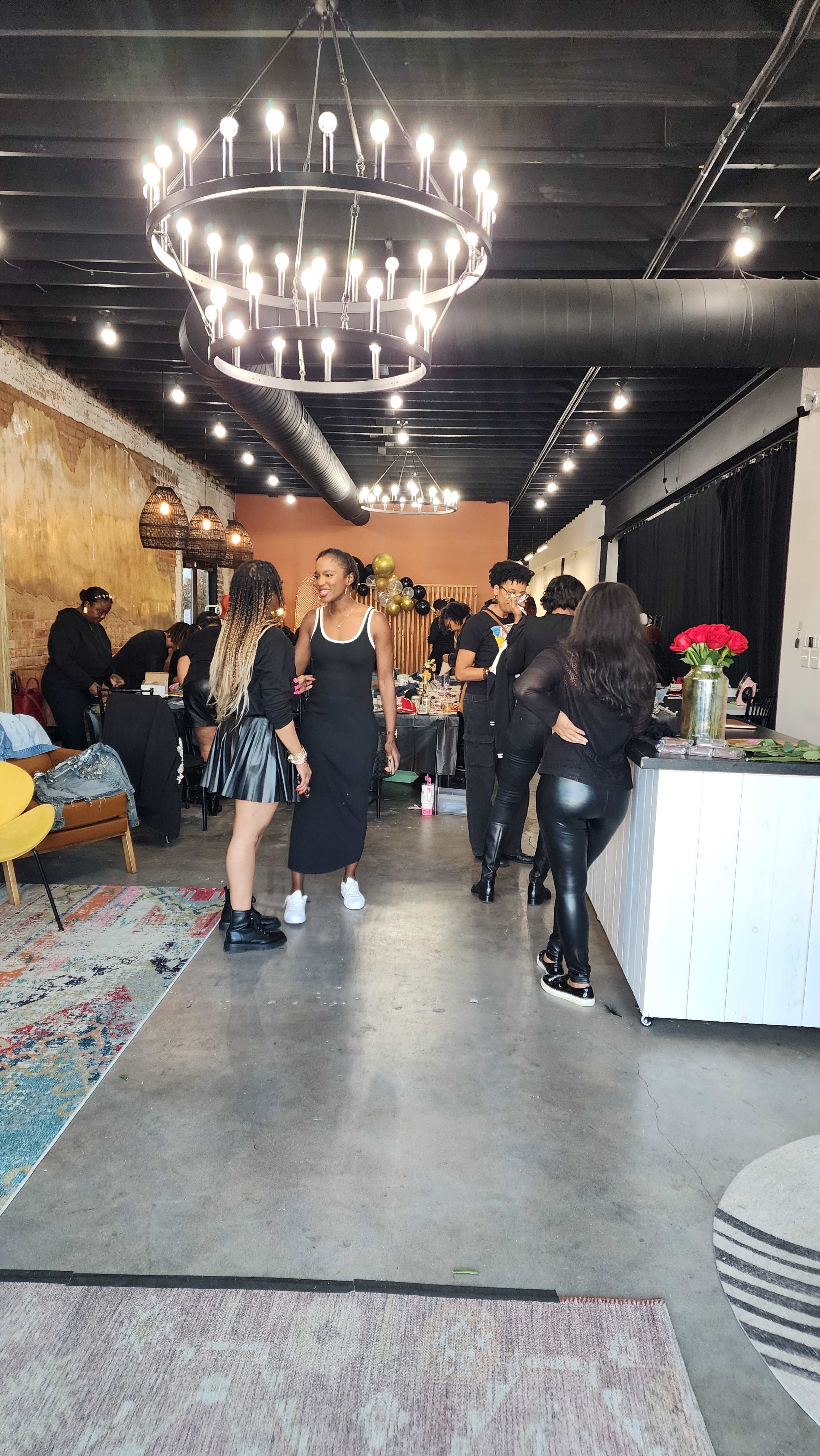 The image shows a modern venue with a chandelier overhead, attendees in black attire socializing, and a display of flowers on the counter.