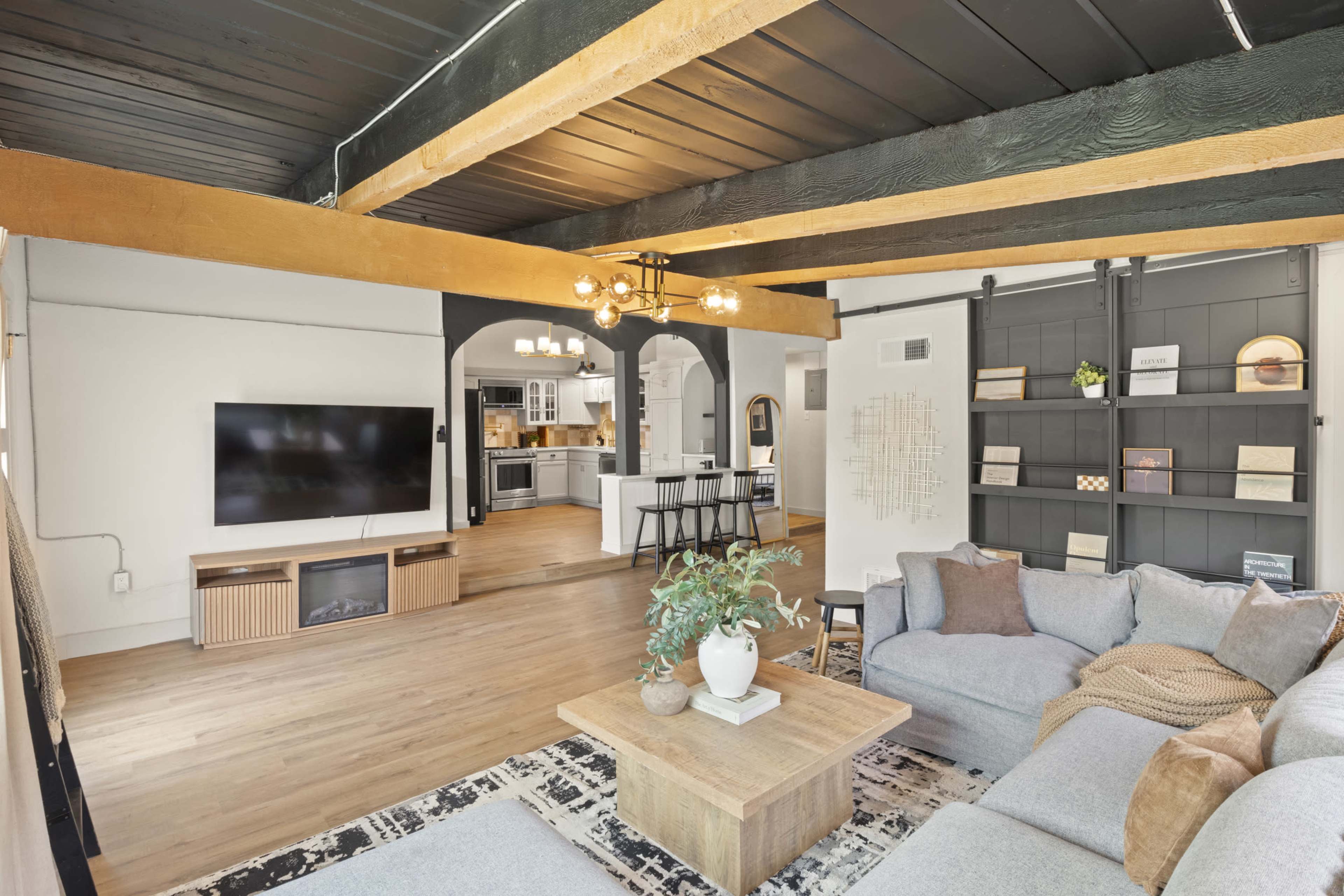 The image shows a modern living room featuring a large sectional sofa, a wooden coffee table, and a mounted television, with a view into a kitchen and dining area beyond.