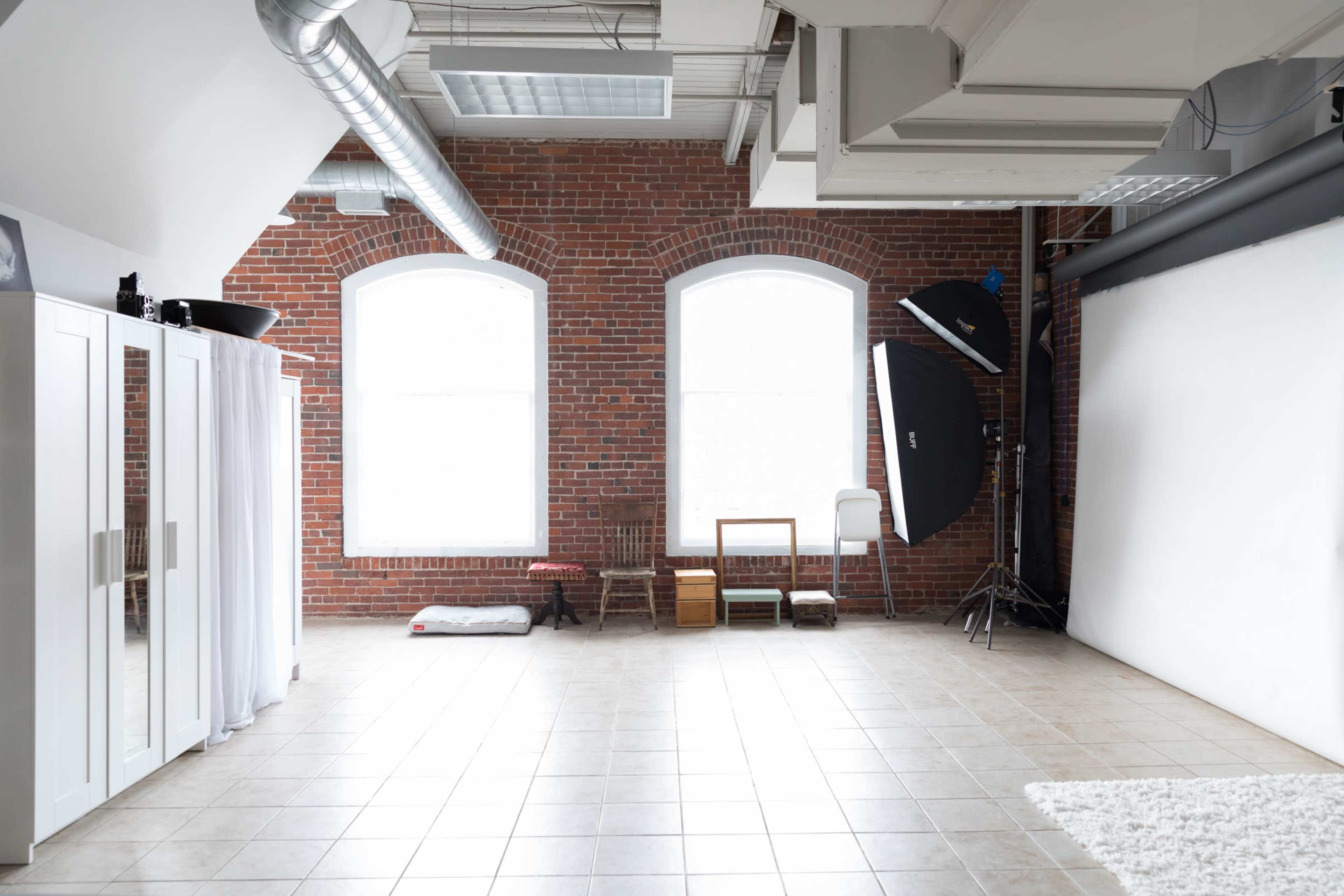 A spacious photography studio with brick walls, large windows, and a backdrop setup, including lighting equipment and props.