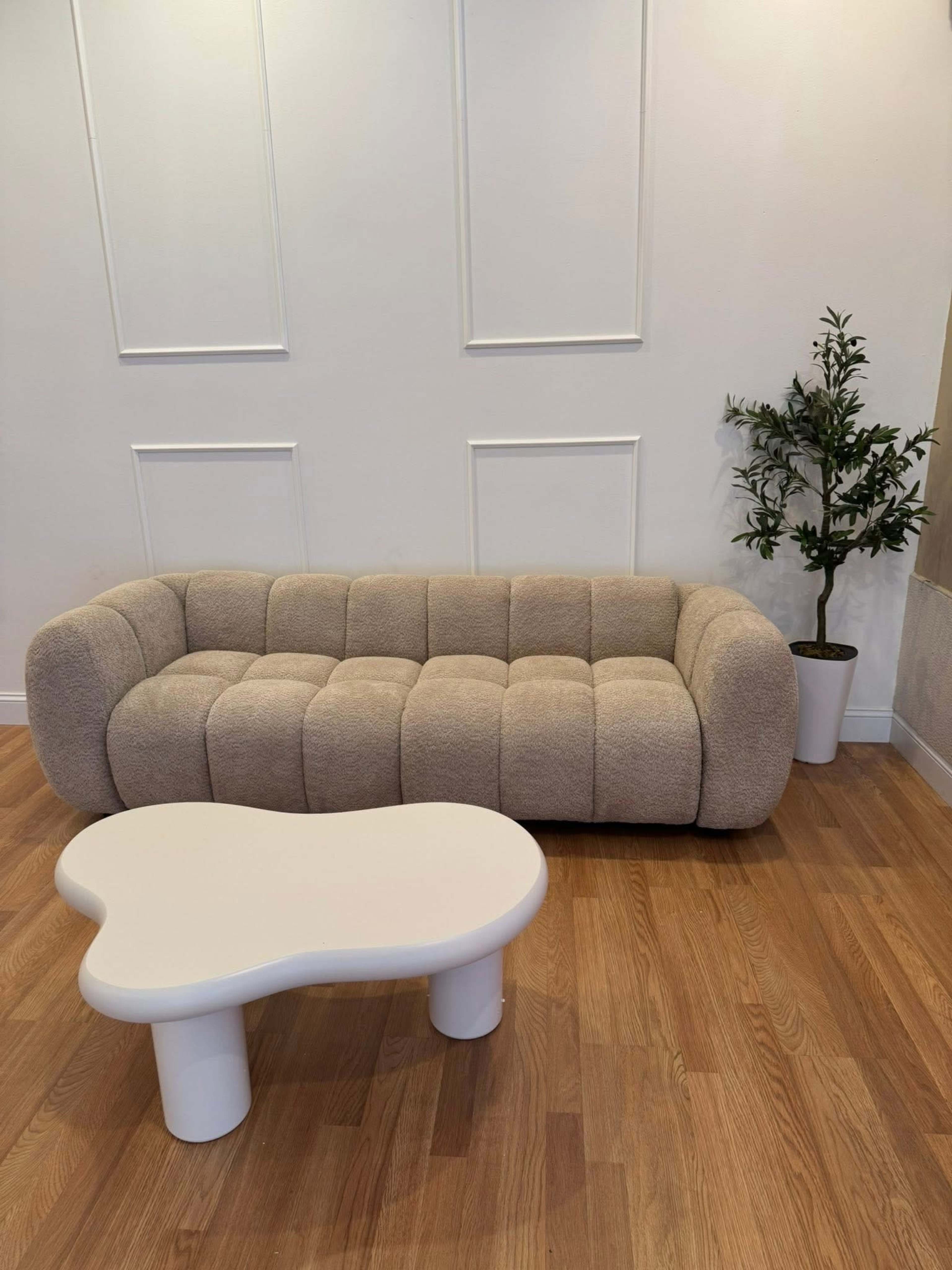 The image shows a light beige, tufted couch next to a white, abstract-shaped coffee table and a small potted plant in a simple white pot, all set against a plain wall with vertical paneling.