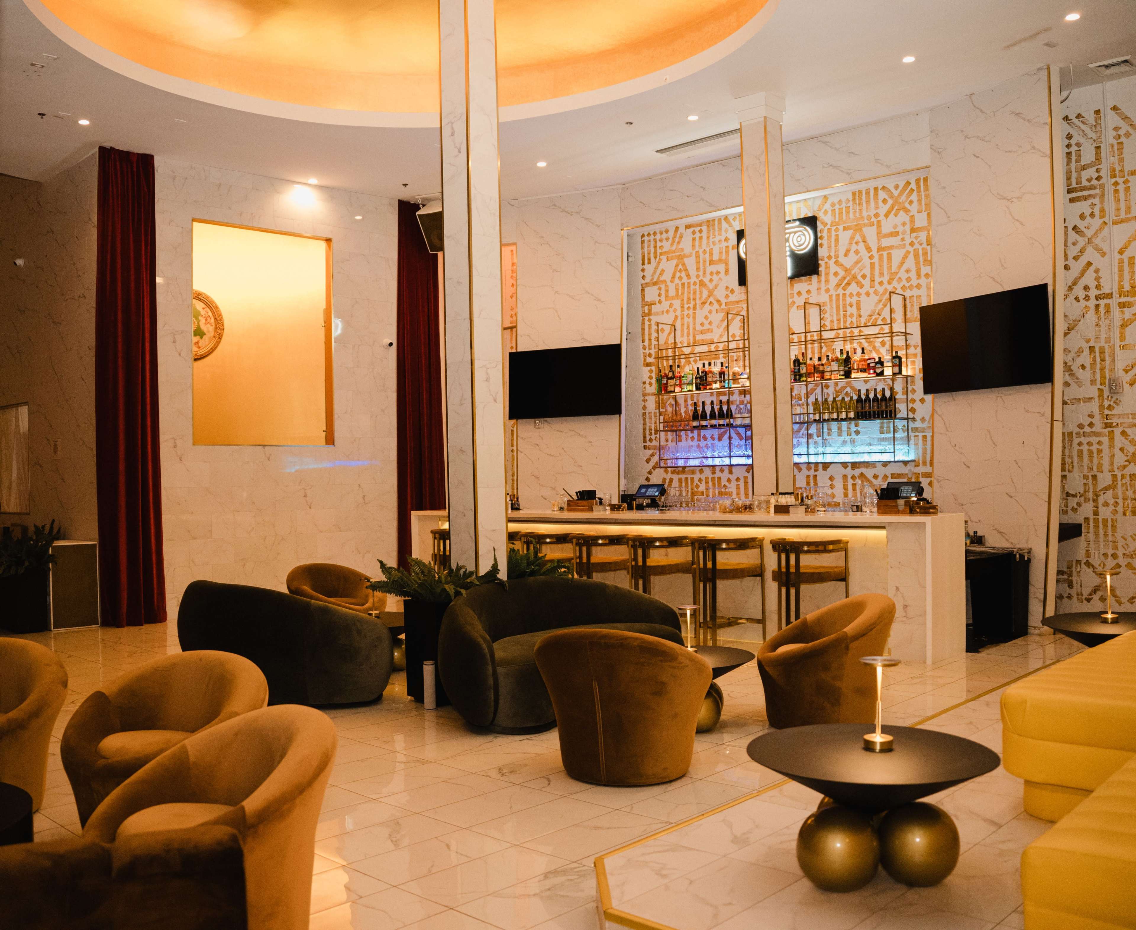 The image shows a modern lounge with a bar, featuring plush seating arrangements and marble accents.