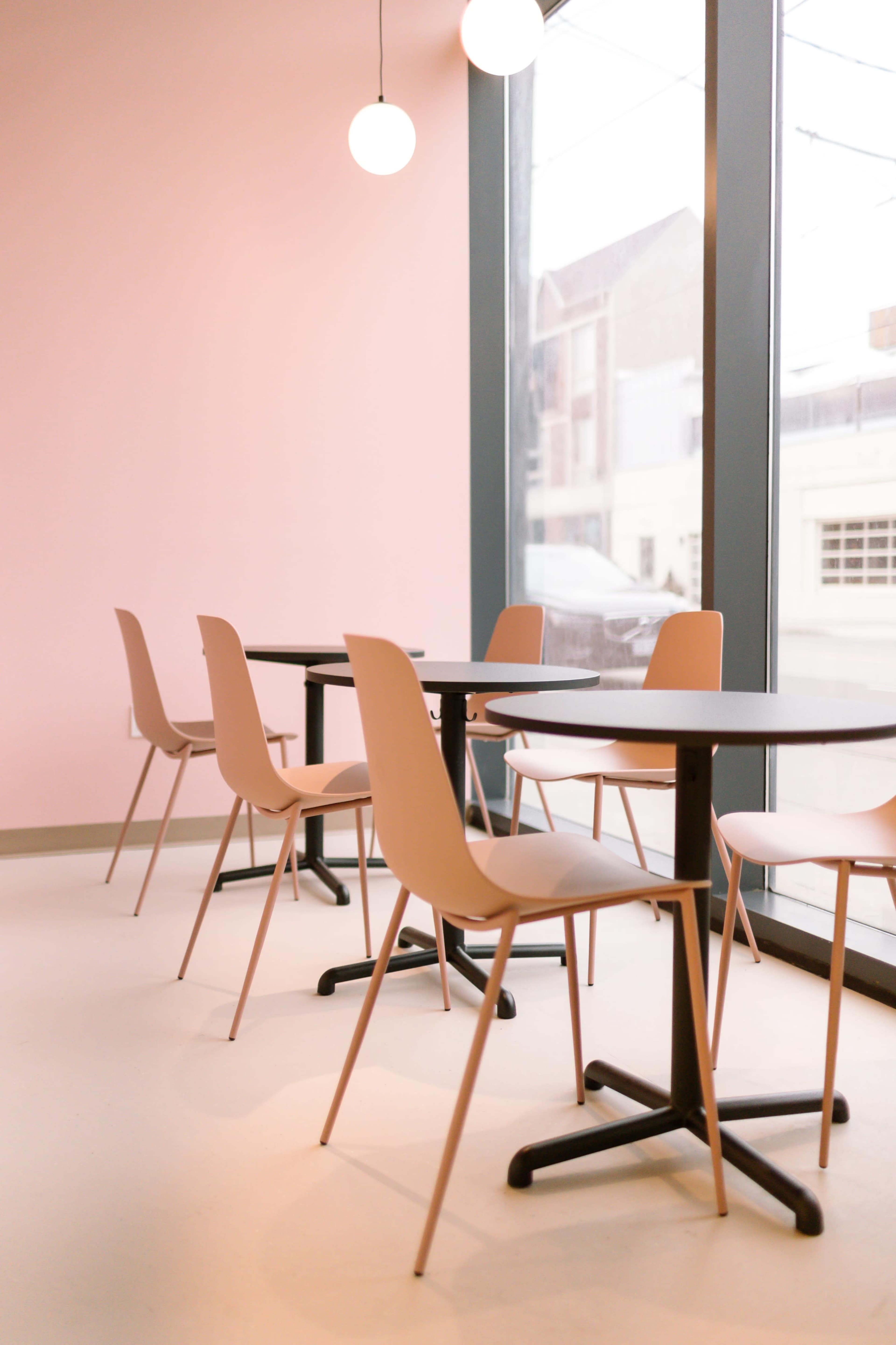 A modern café interior features pink walls, round pendant lights, and minimalist tables paired with chairs arranged around large windows.