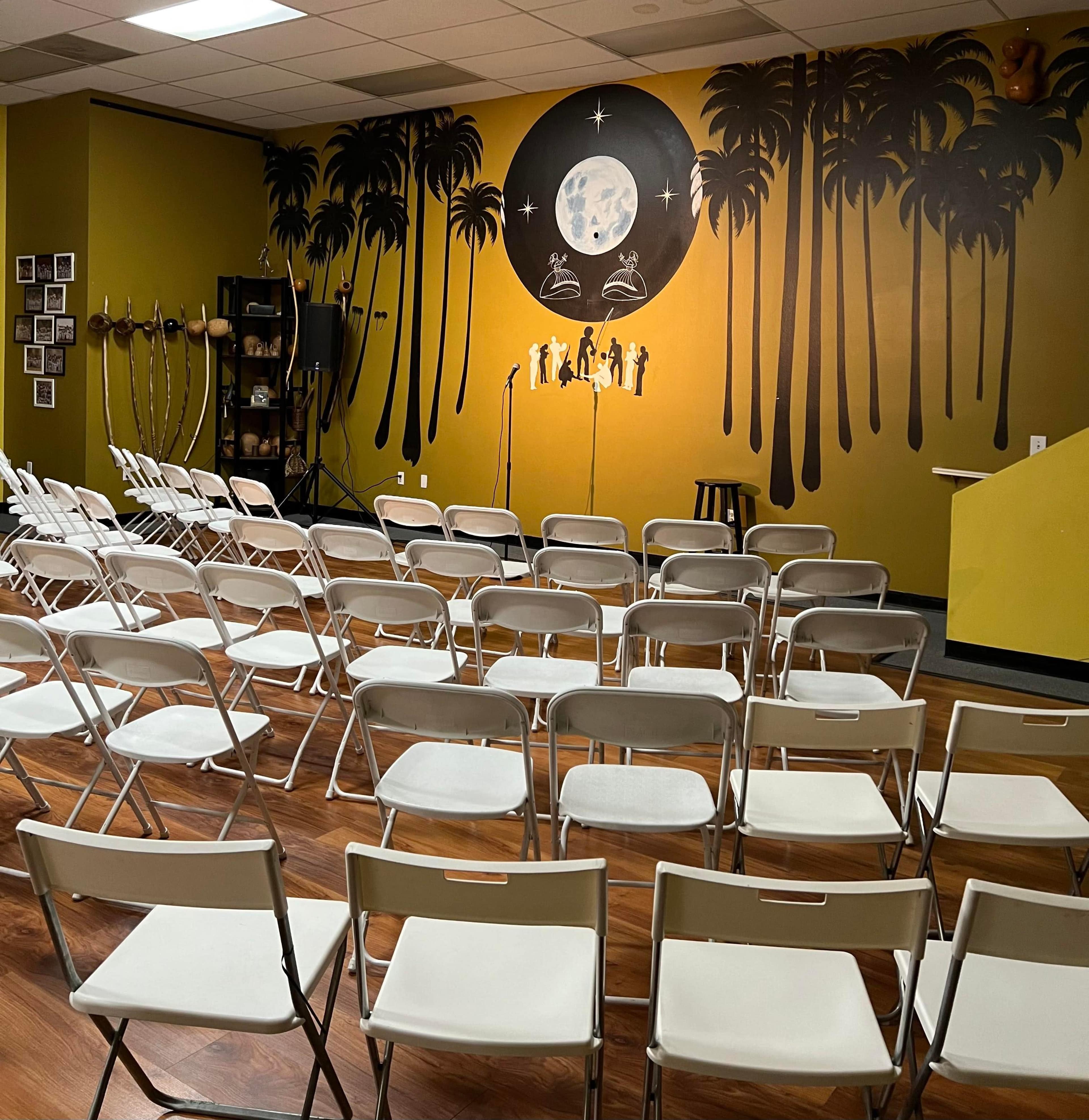 A room set up for a performance, featuring rows of white folding chairs facing a stage with microphones in front of a mural of palm trees and a moon.