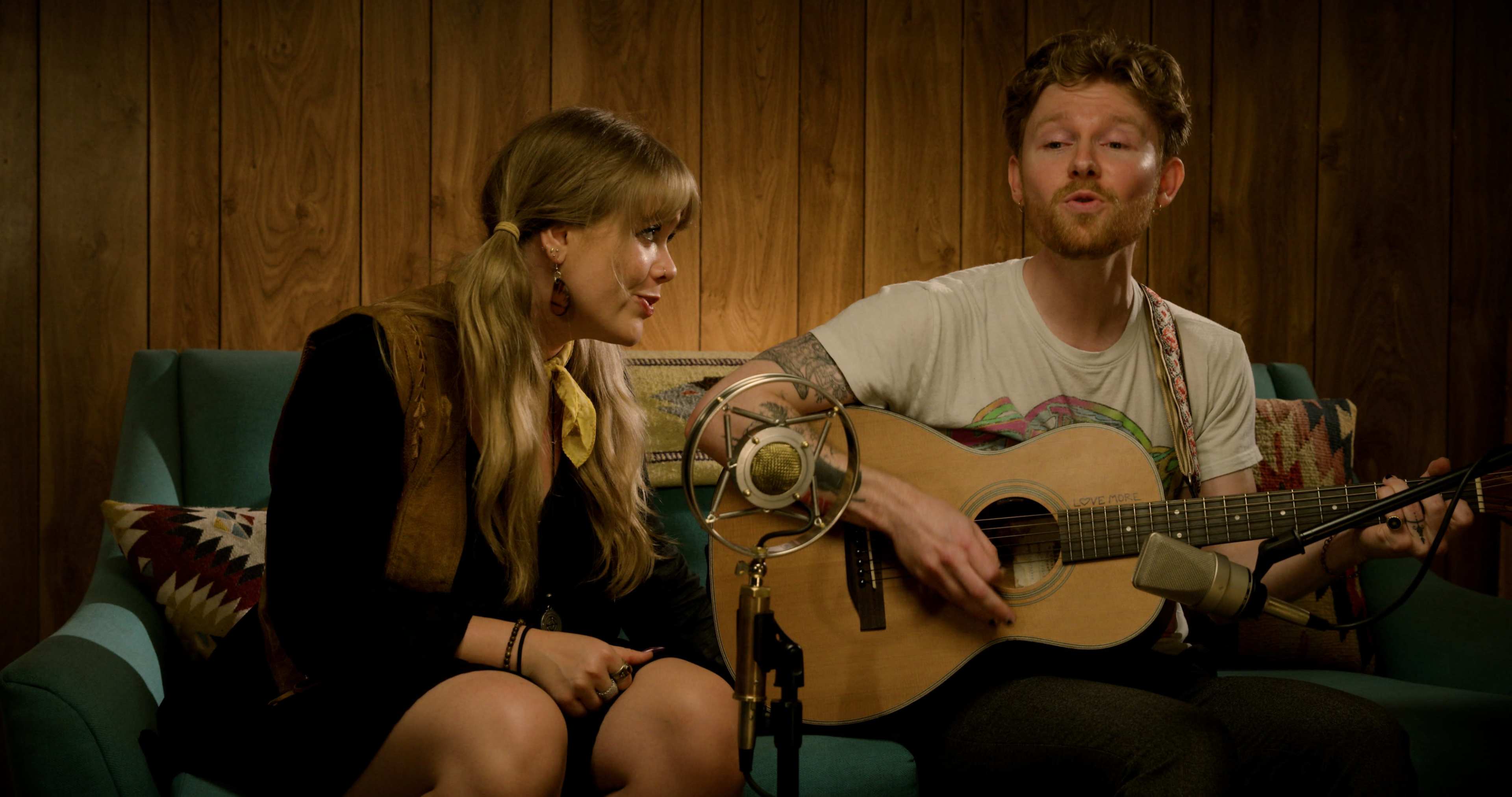 A man plays an acoustic guitar and sings while a woman listens intently in front of a wooden wall.
