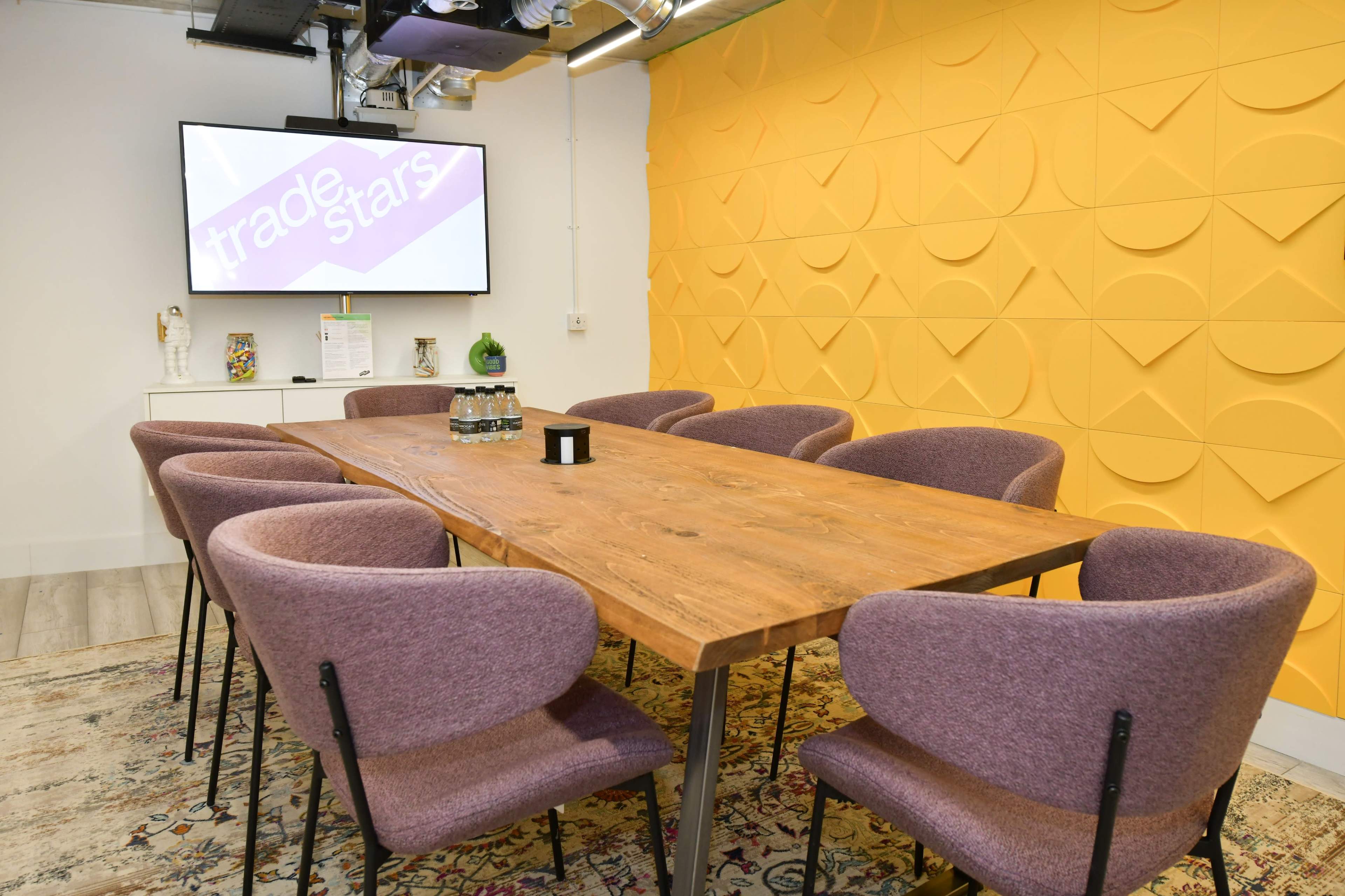 A modern meeting room features a large wooden table surrounded by purple chairs, with a screen displaying the text "trade stars" on a yellow wall.