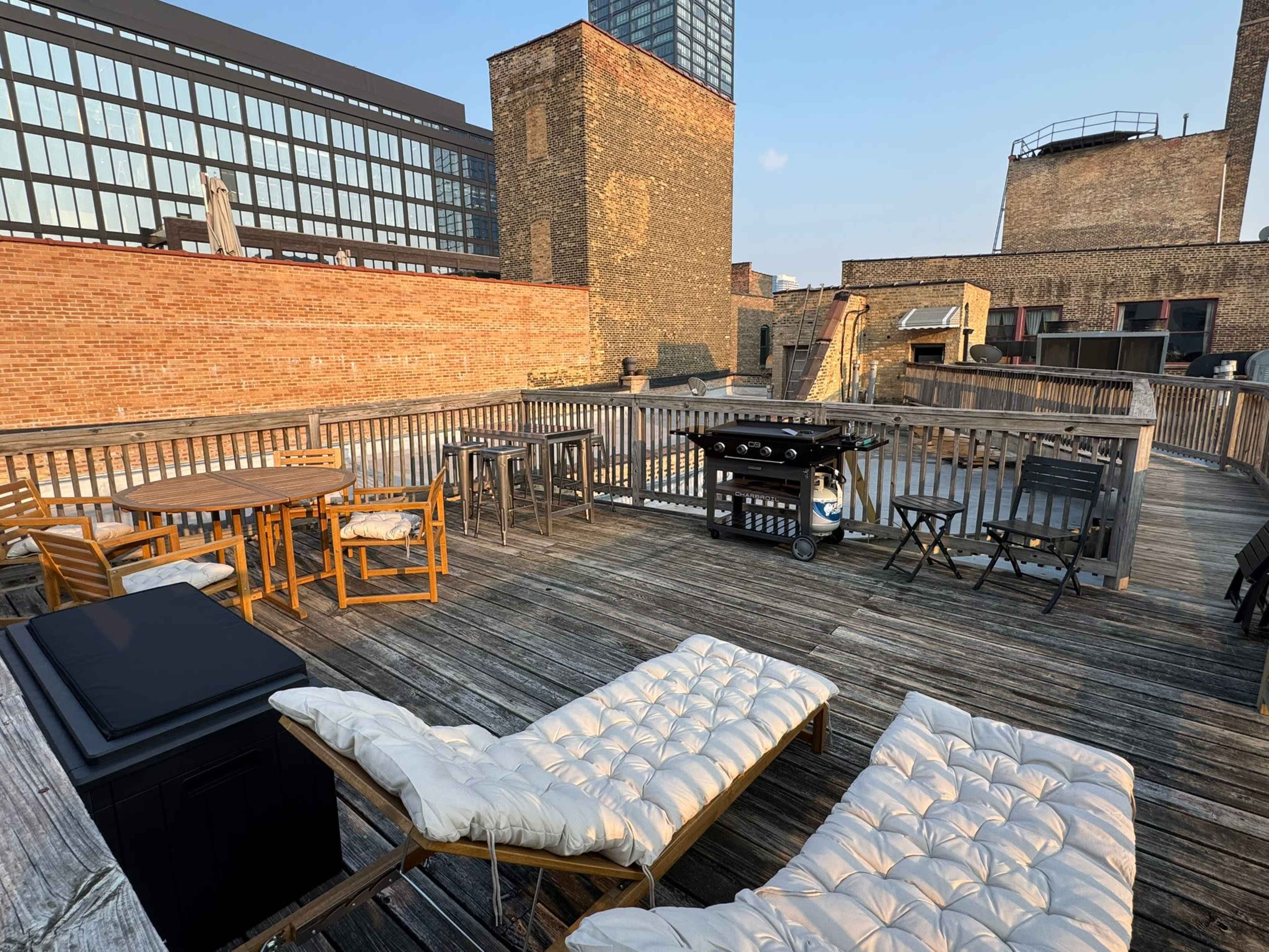 The image shows a rooftop deck with wooden flooring, featuring lounge chairs, tables, a grill, and a view of surrounding buildings.