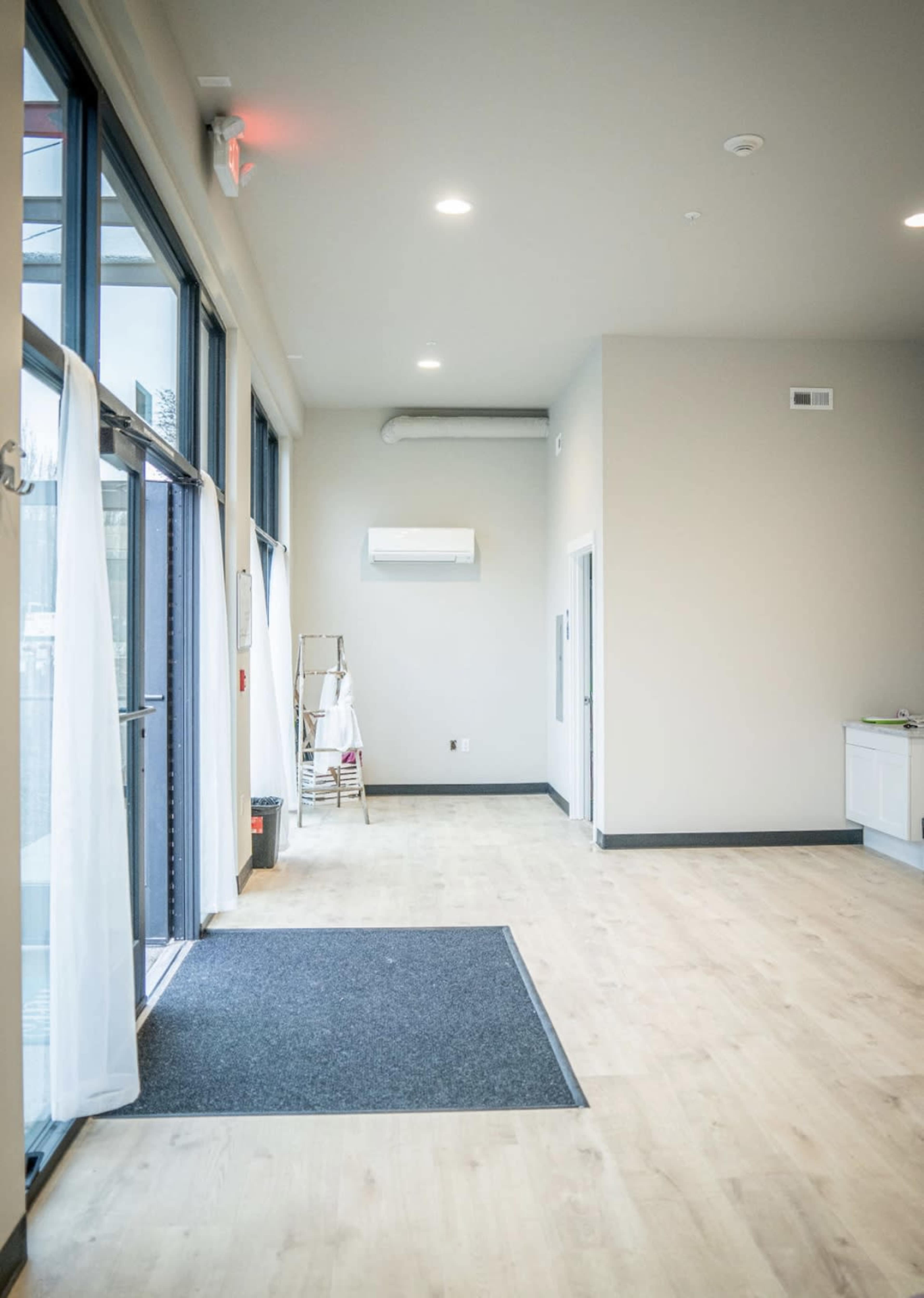 The image shows a spacious, empty room with large windows, light-colored walls, and a gray carpet mat at the entrance.
