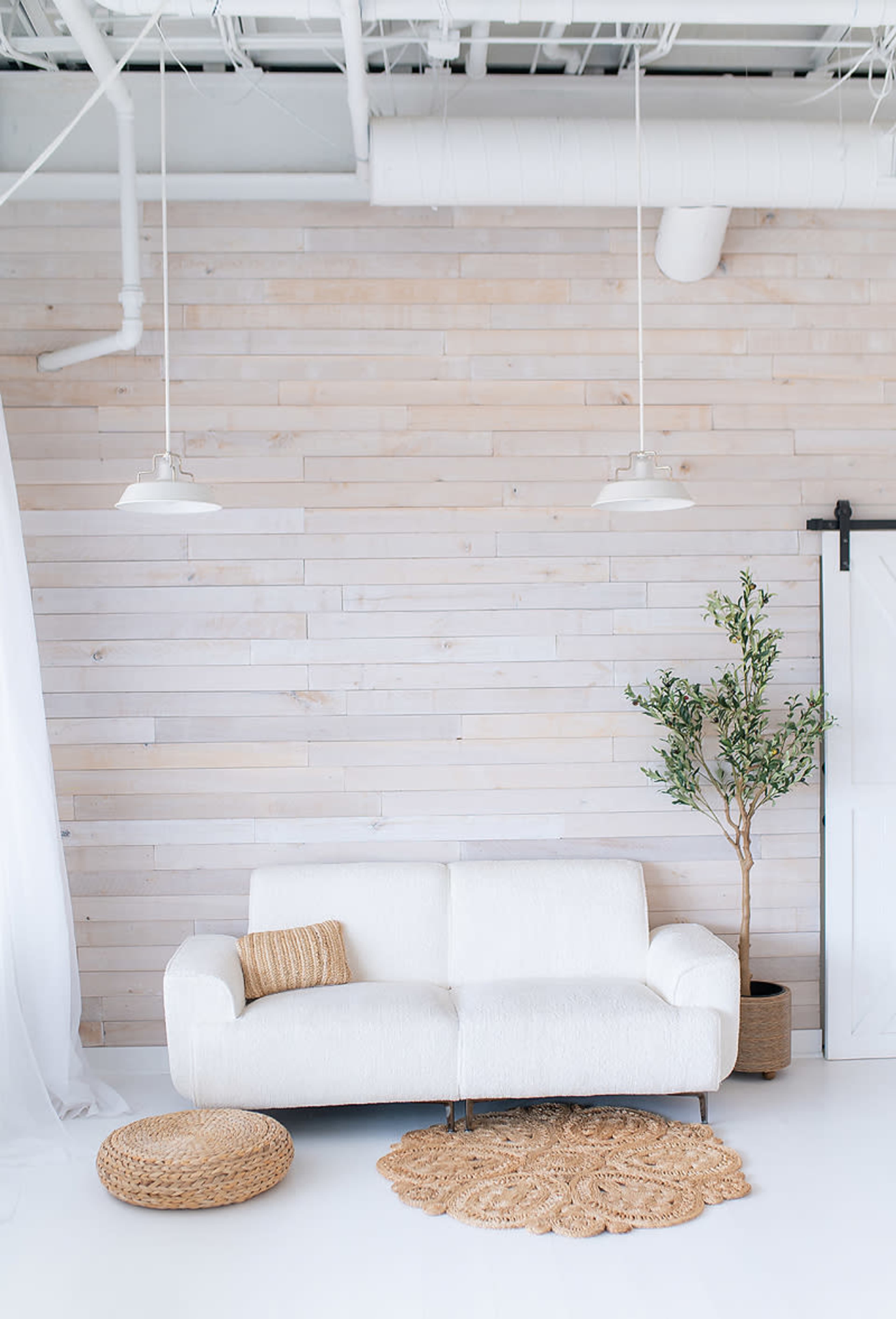 A white sofa sits against a light wooden wall, accompanied by a potted plant and woven decor on the floor.