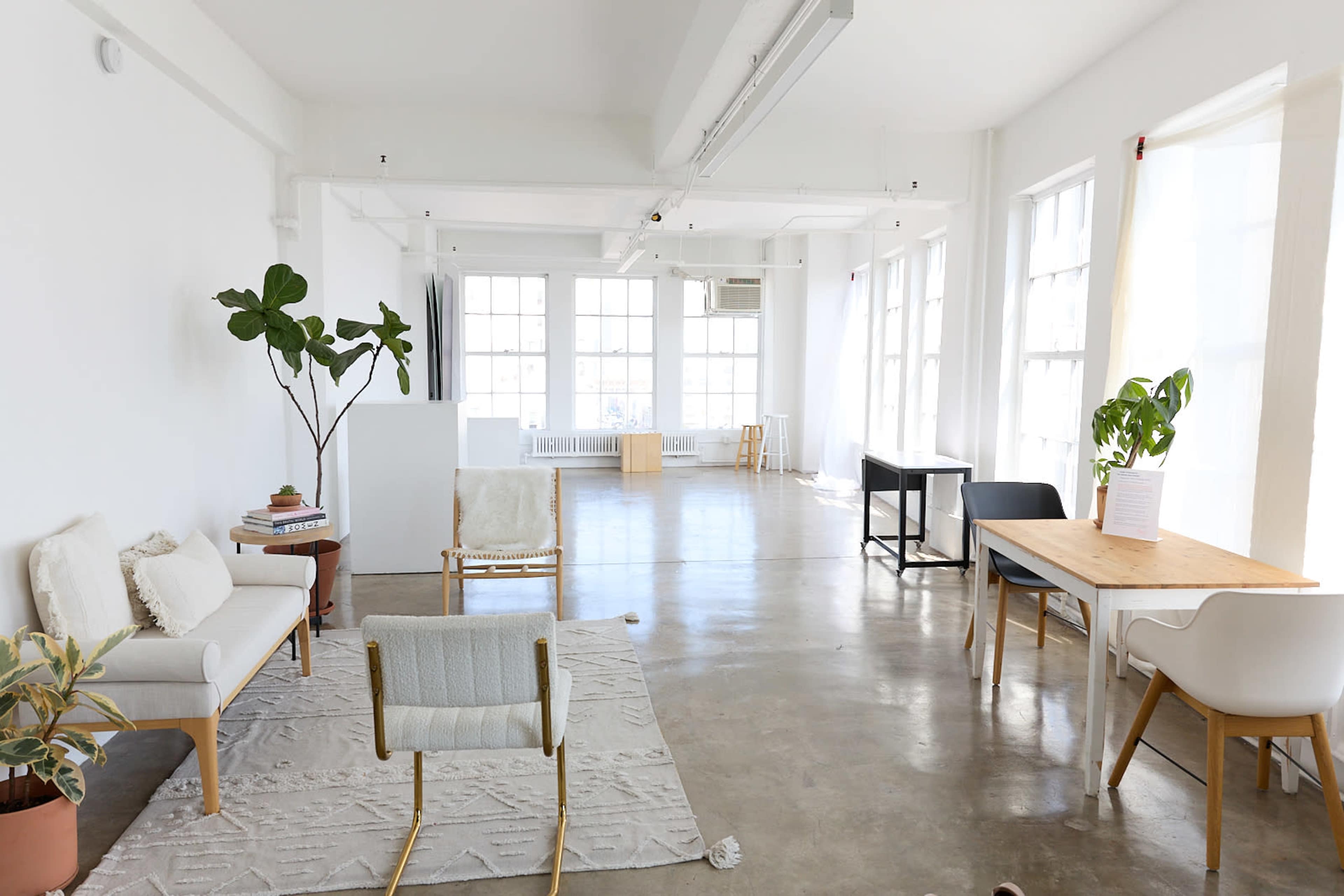 A bright, spacious room with large windows, minimalist furniture, and a neutral color palette, featuring seating areas and a wooden table.