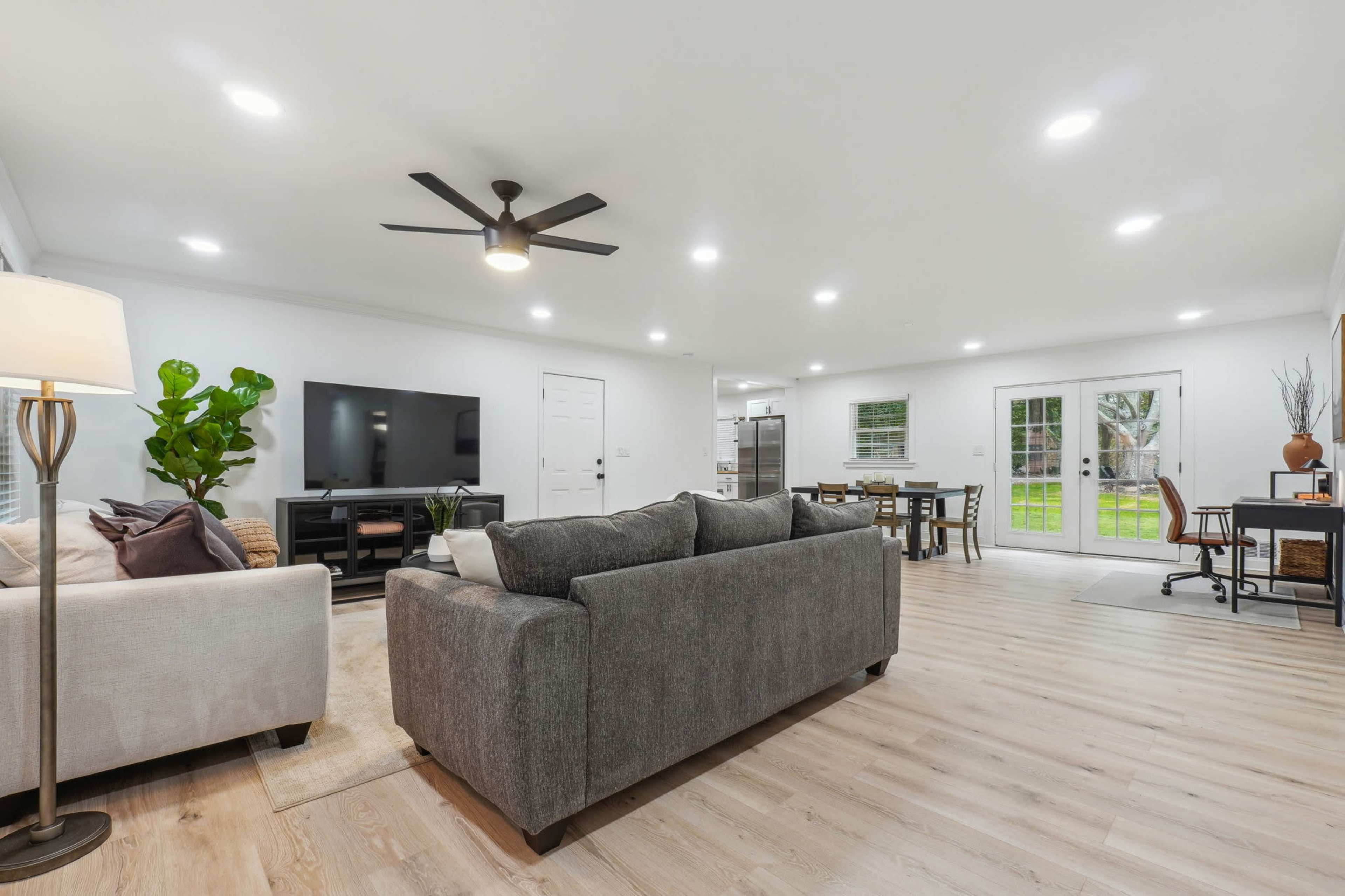 A modern living area featuring a gray sofa, a light-colored armchair, a television stand, and a dining table with chairs, all set in a well-lit space with sliding glass doors leading to an outdoor area.