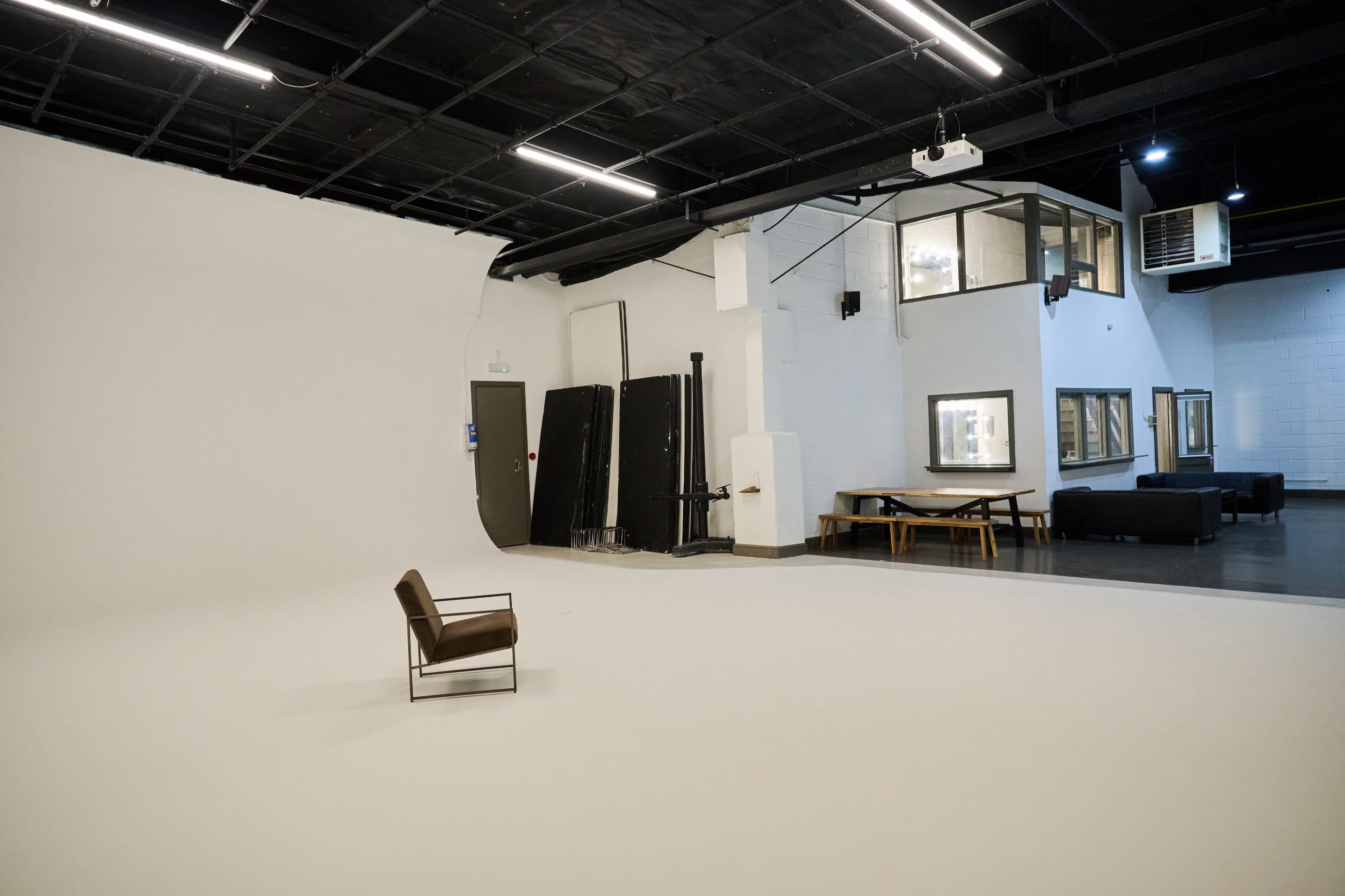 The image shows an empty studio space featuring a single chair on a white floor, with a backdrop of large windows and furniture along the walls.