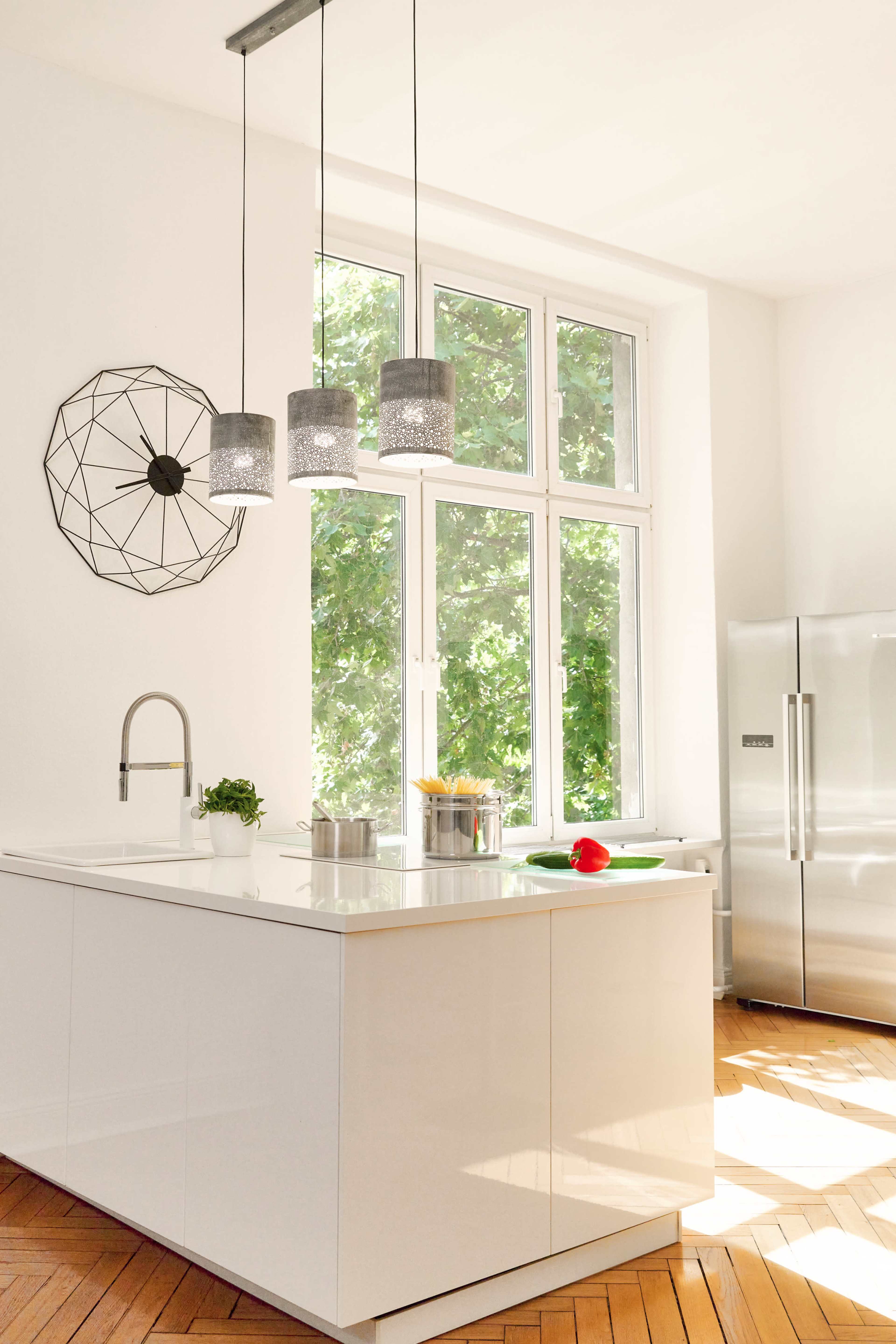 A modern kitchen features a central island with three pendant lights, large windows allowing ample natural light, and stainless steel appliances.