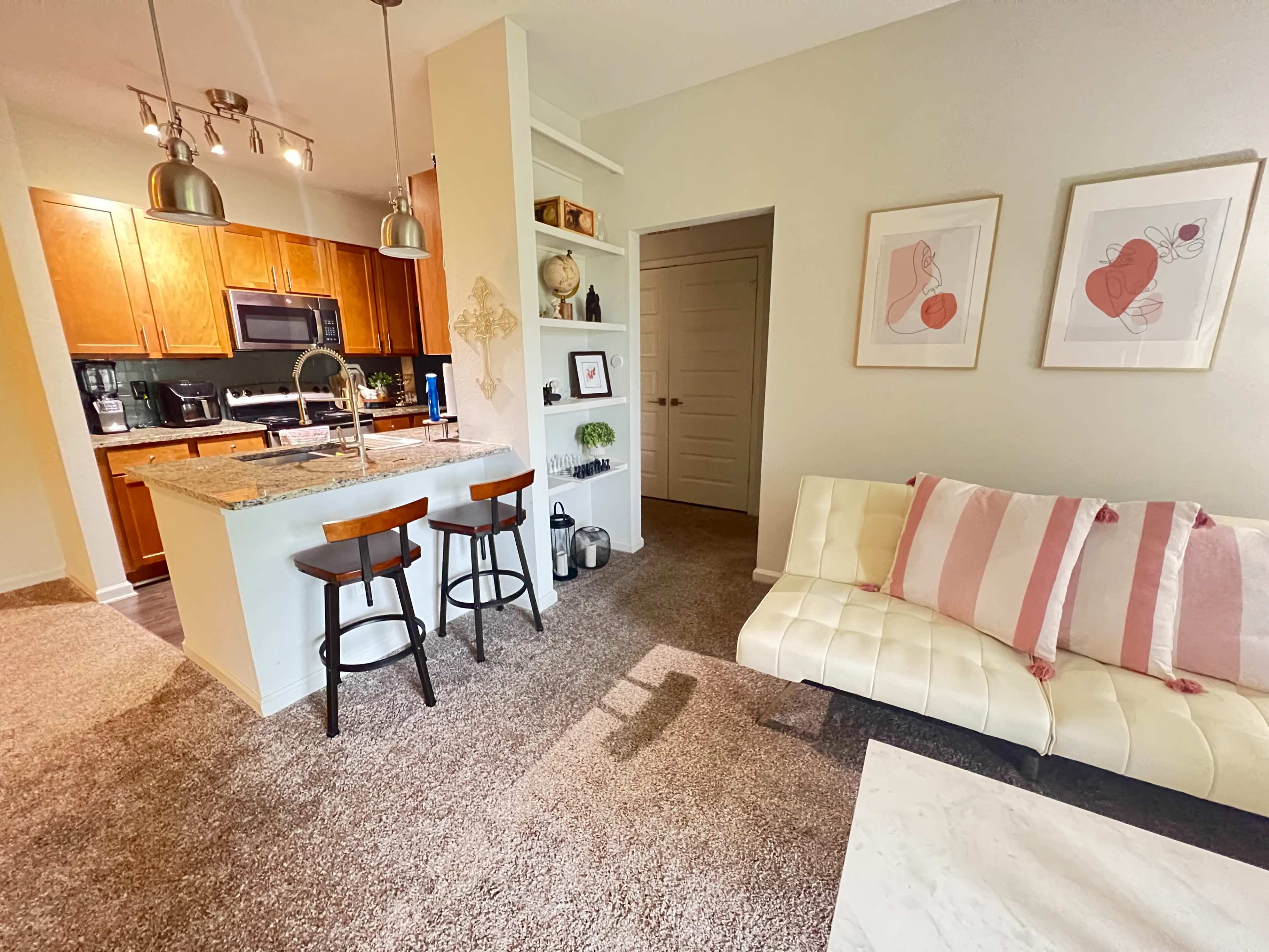 A cozy living area features a white couch with pink striped pillows, an adjacent kitchen with wooden cabinets, and a small dining space with two bar stools.