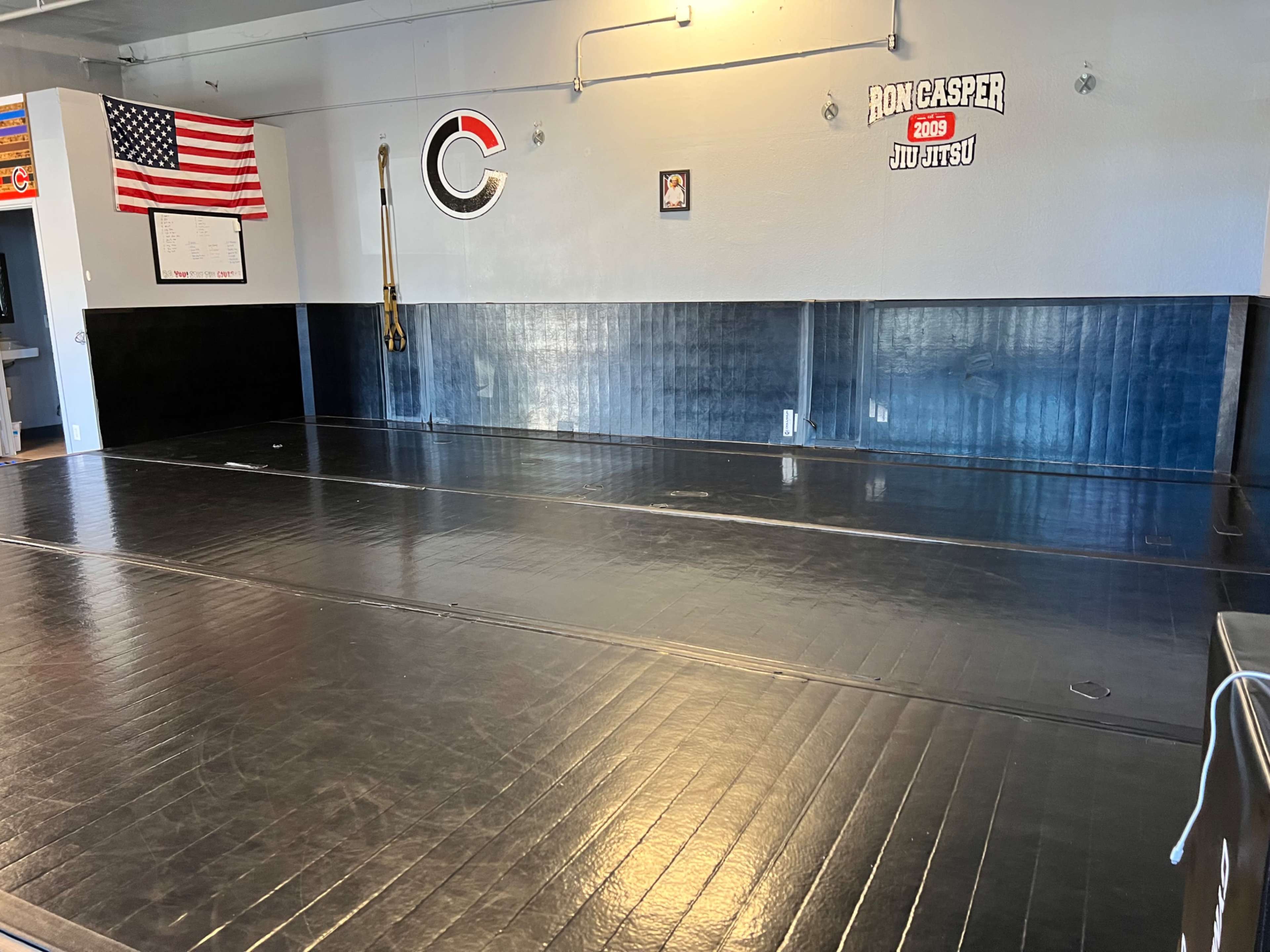 The image shows a spacious jiu-jitsu training mat area with black flooring and a wall displaying an American flag and gym branding.