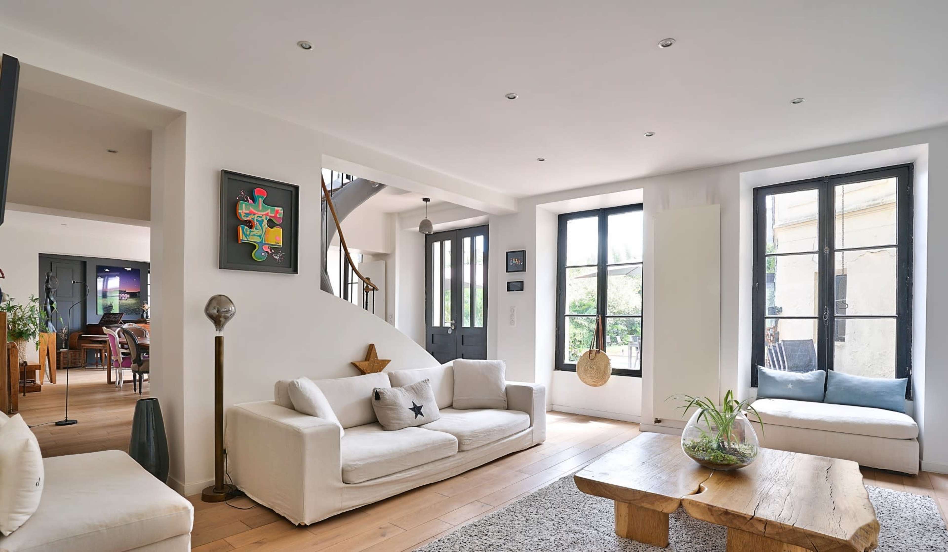 A modern living room with white sofas, a wooden coffee table, and large windows that let in natural light.