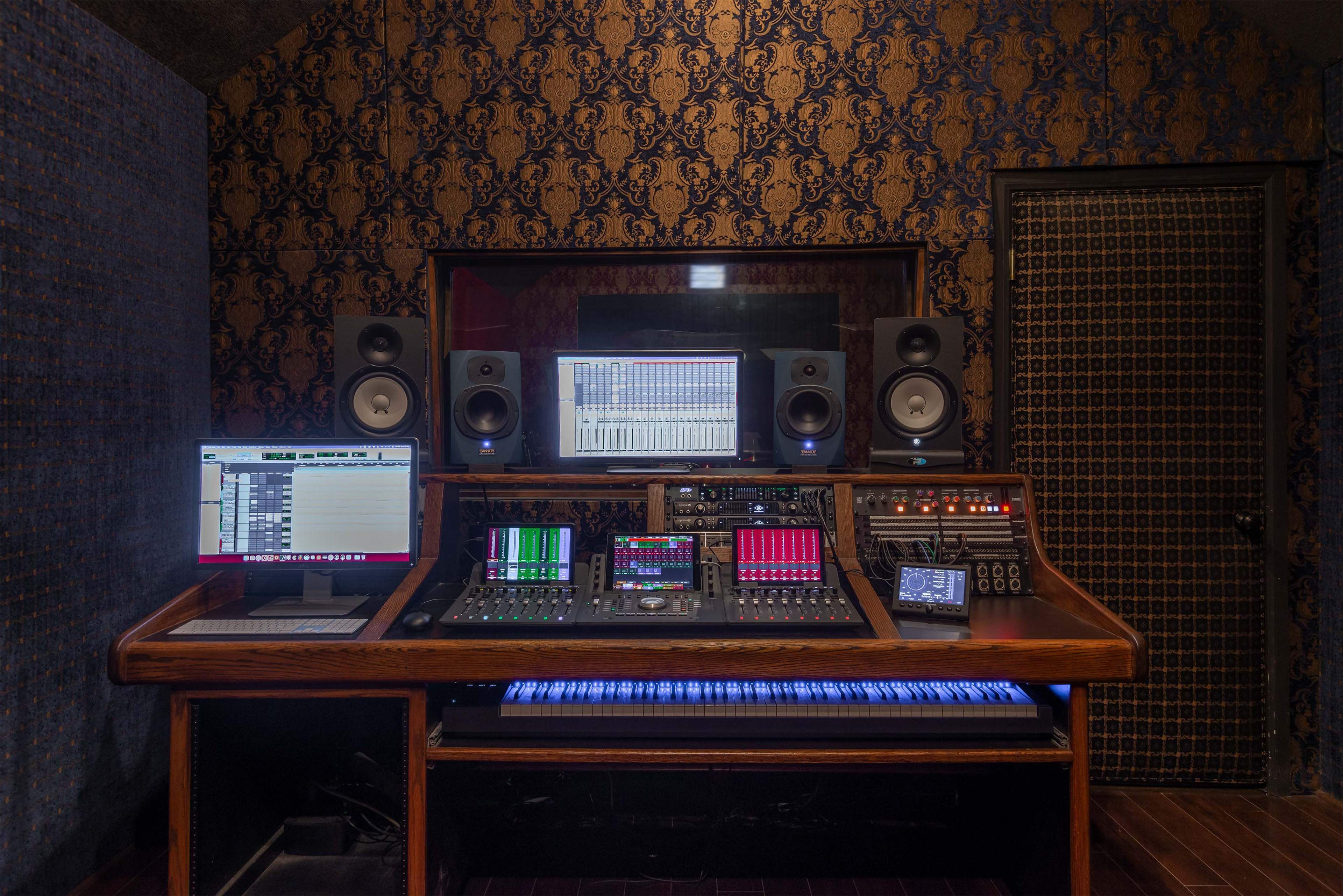 A recording studio is shown with a mixing console, computers, and speakers arranged on a wooden desk against a decorative wall.