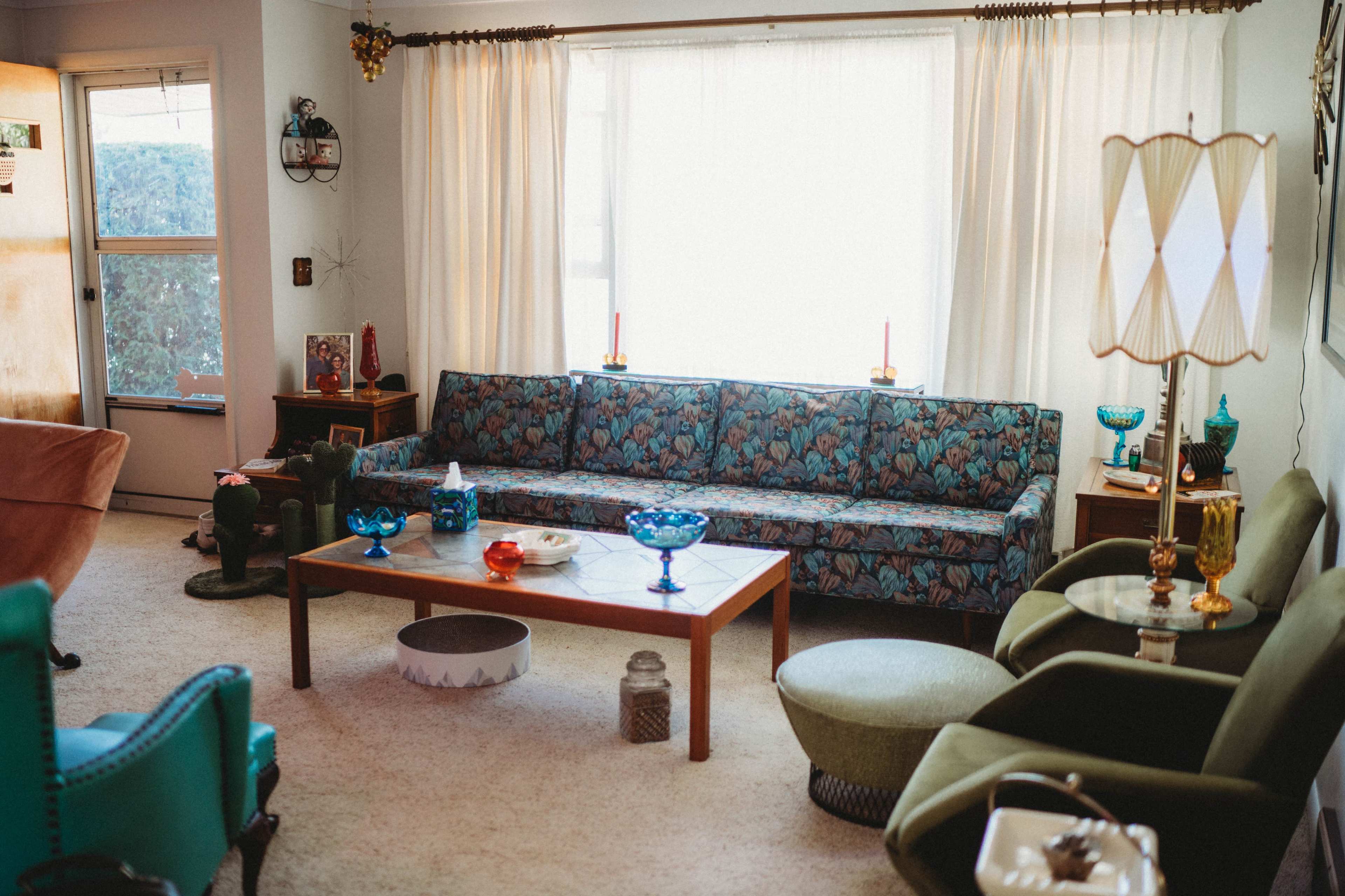 The image shows a vintage living room with a floral-patterned sofa, a wooden coffee table, and various decorative items, including colored glass bowls and a floor lamp.
