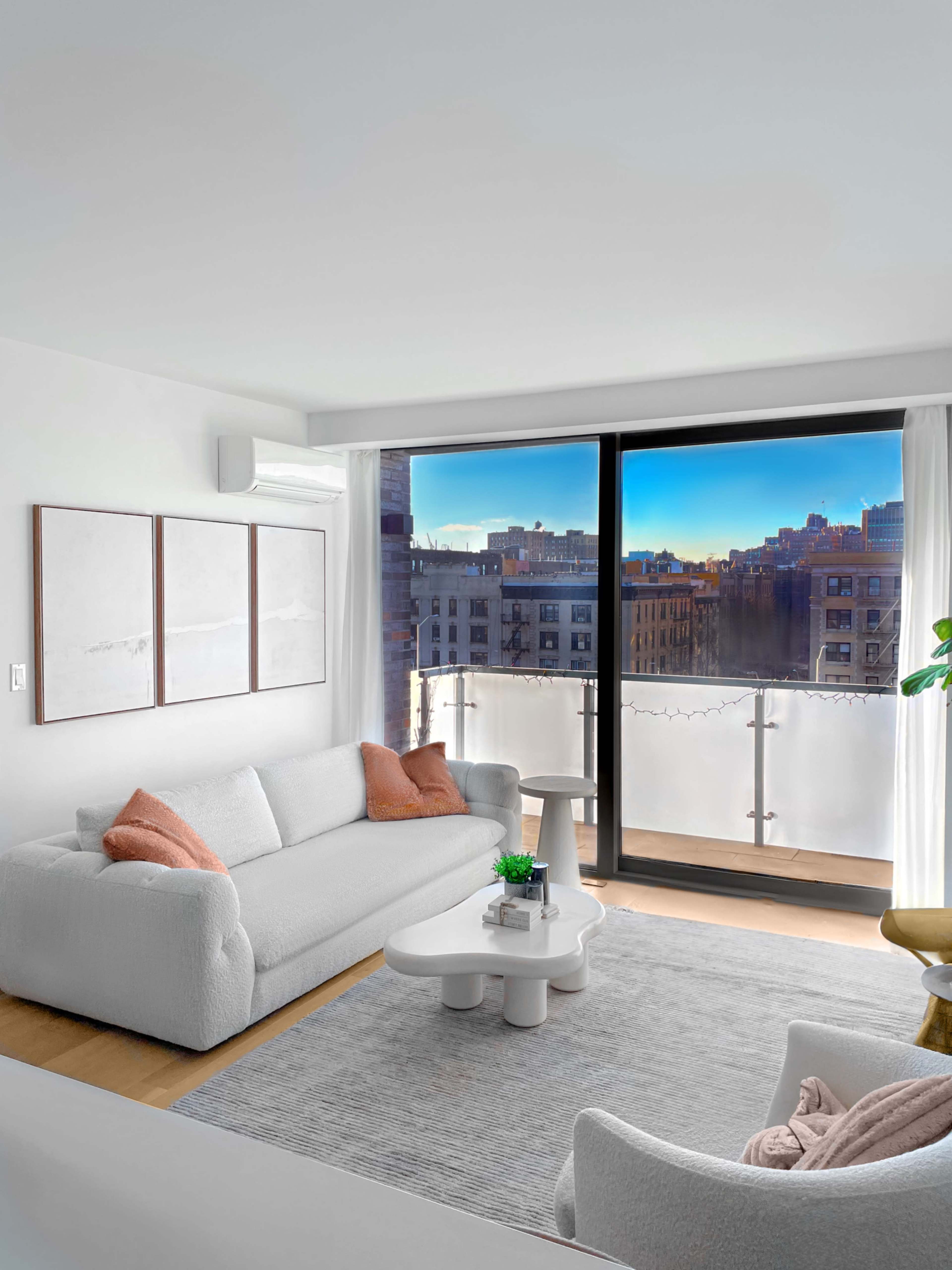A modern living room features a white sofa and armchair, a coffee table, and large windows showcasing an urban view.