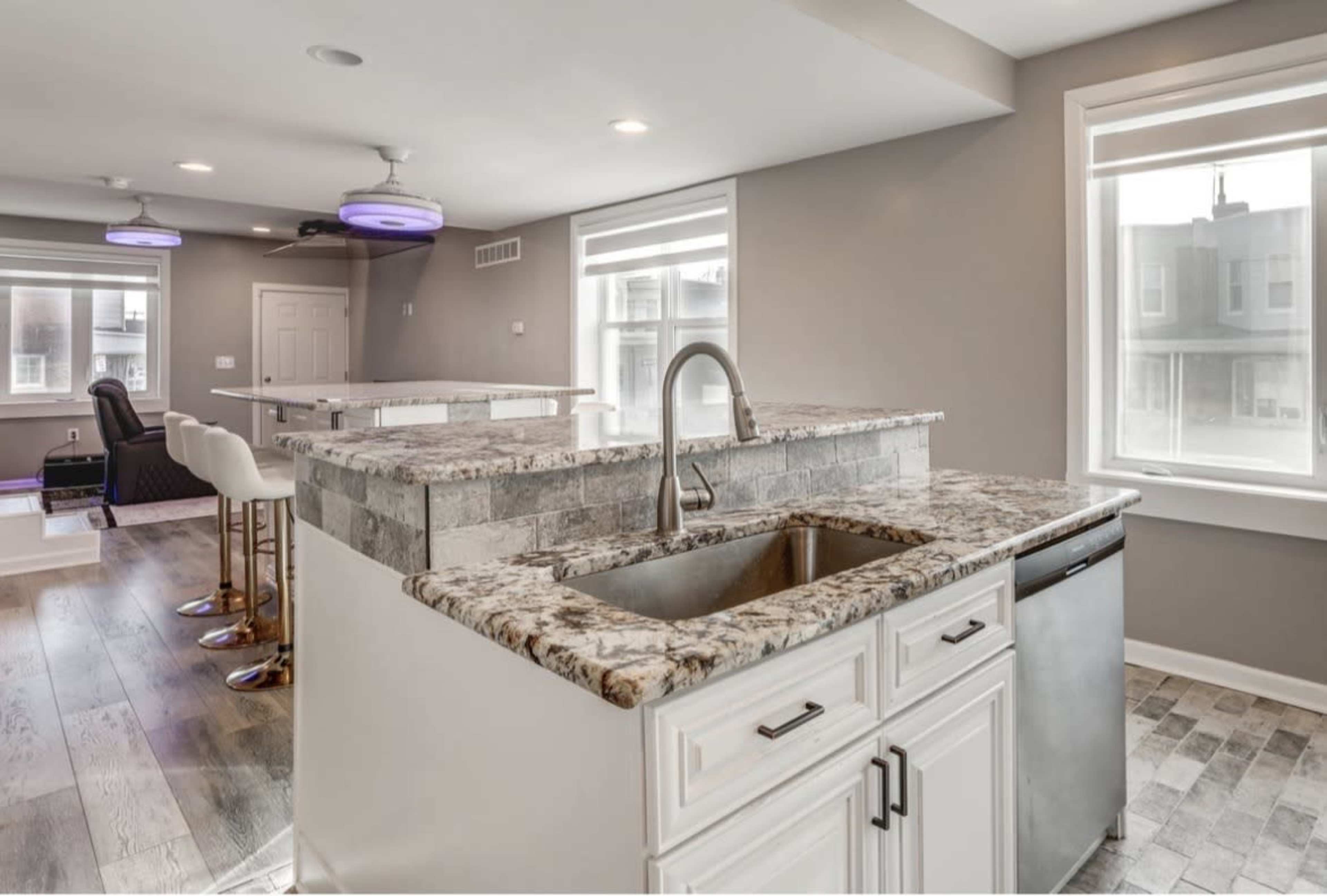 The image shows a modern kitchen featuring a granite countertop with a sink, stainless steel dishwasher, and open space with large windows letting in natural light.