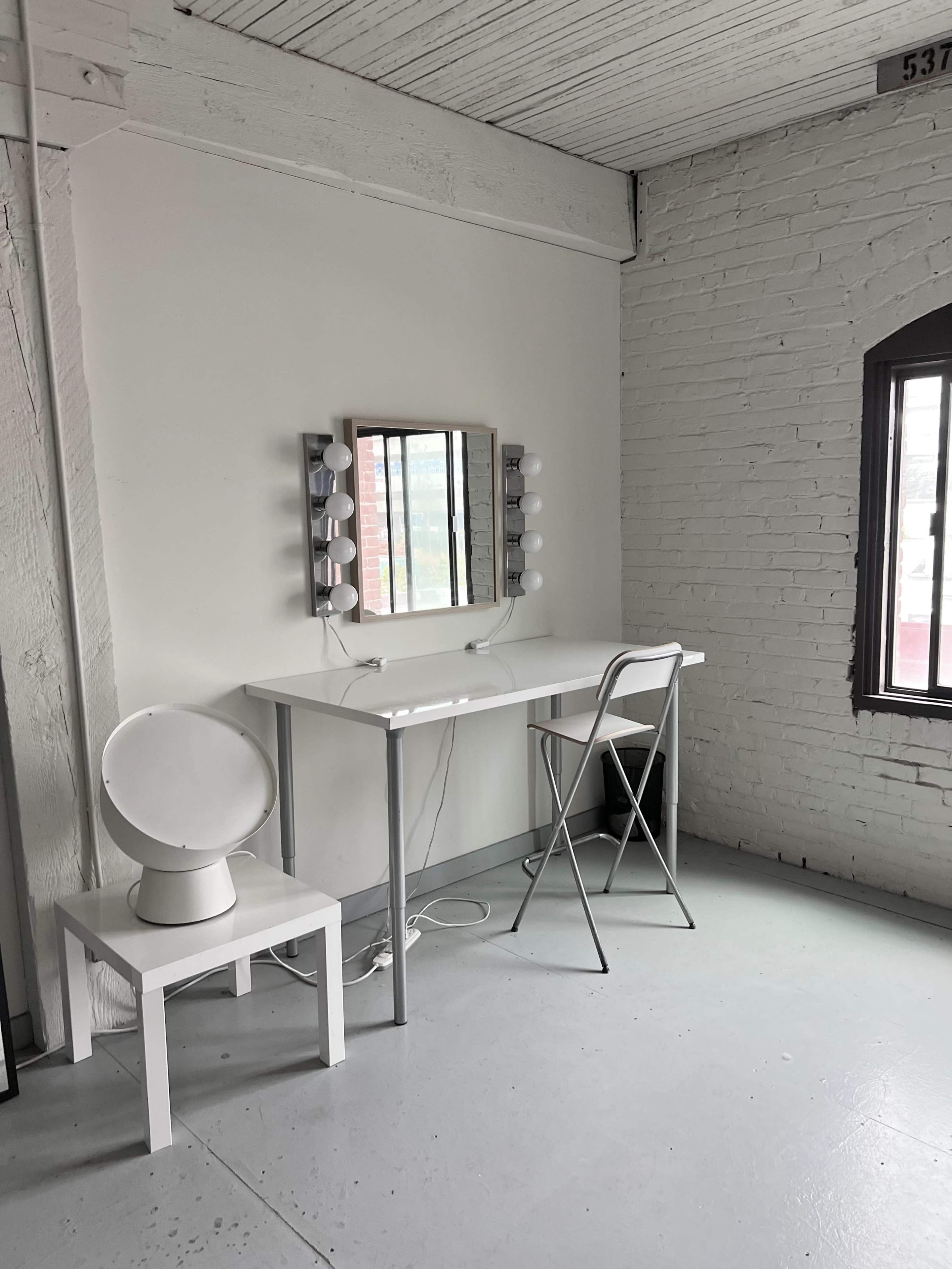 A minimalist makeup vanity with a mirror and lights sits against a white brick wall beside a chair and circular stool in a well-lit room.
