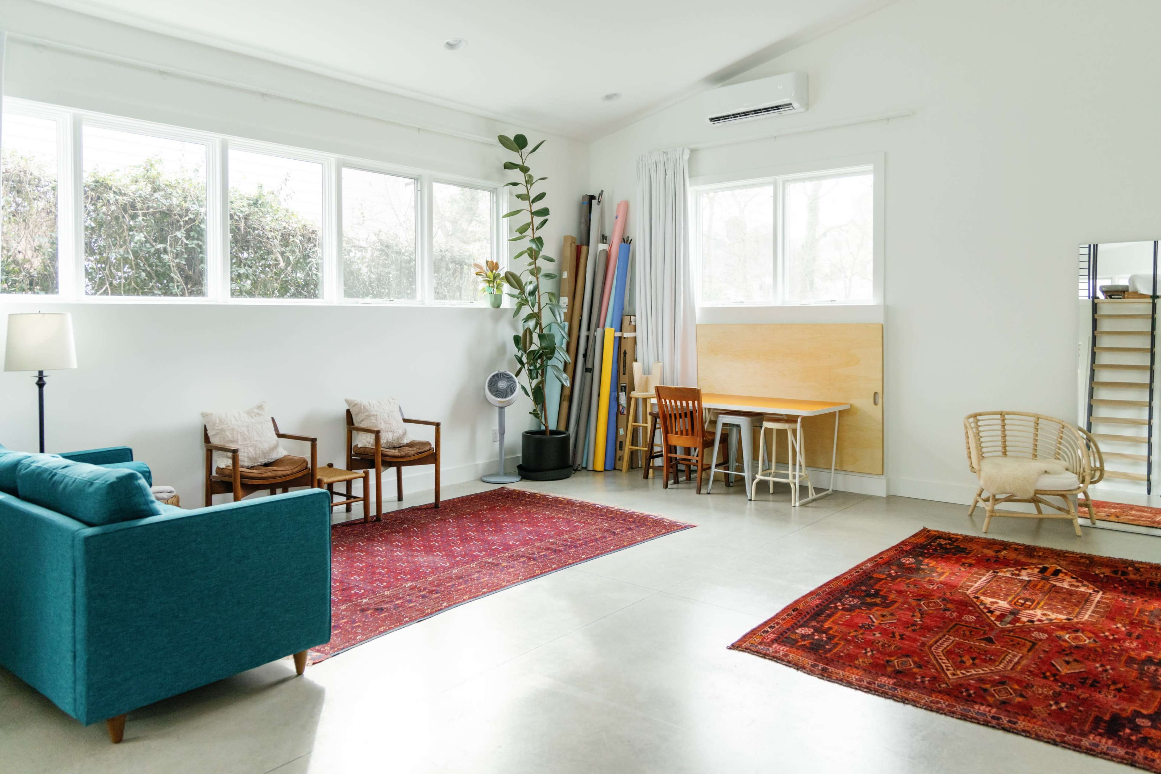 A bright, spacious room features a teal sofa, wooden chairs, a table, and decorative rugs against a backdrop of large windows and light-colored walls.