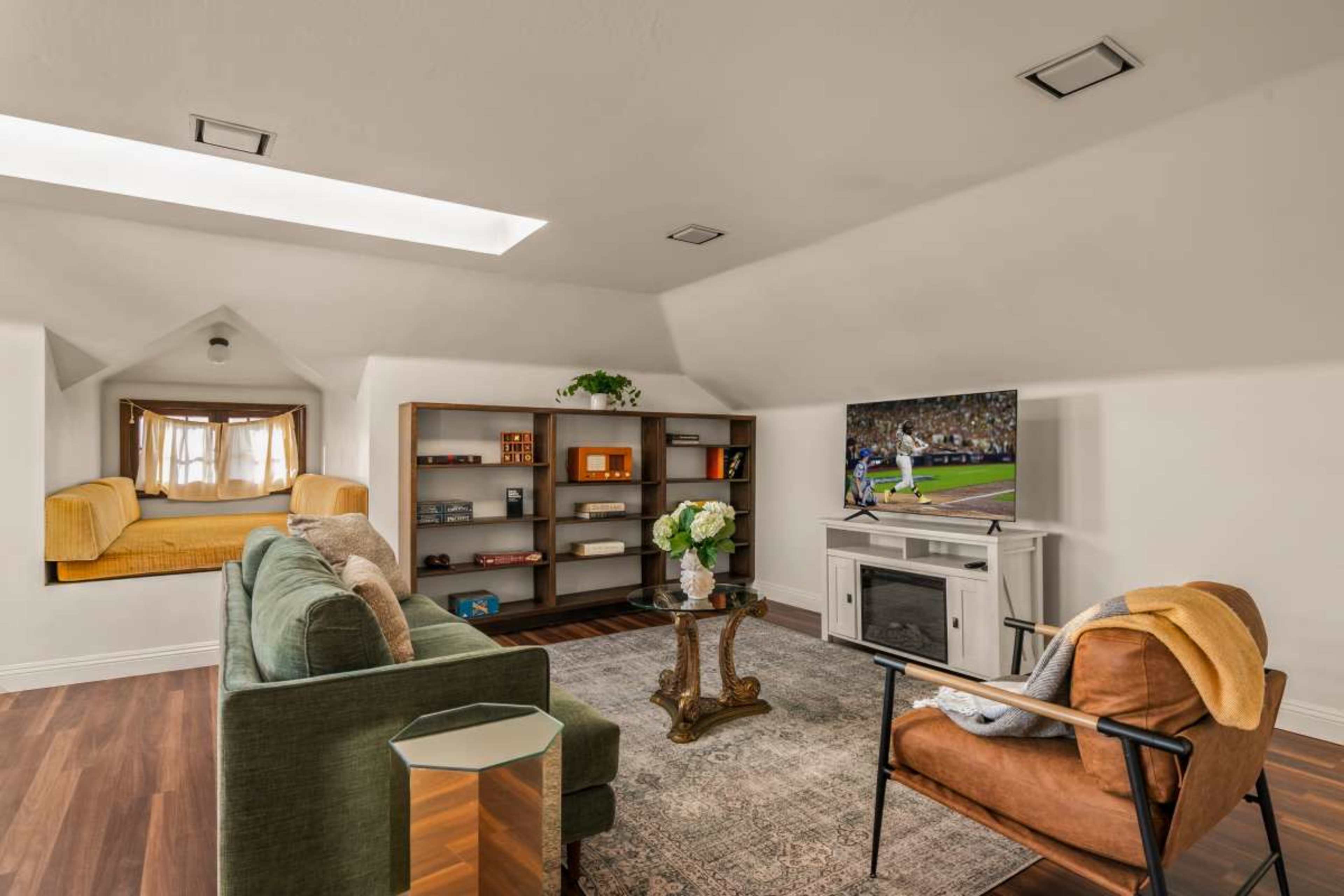 The image shows an attic room furnished with a green sofa, an orange armchair, a coffee table, and a television mounted on a white entertainment unit, all arranged around a decorative area rug.
