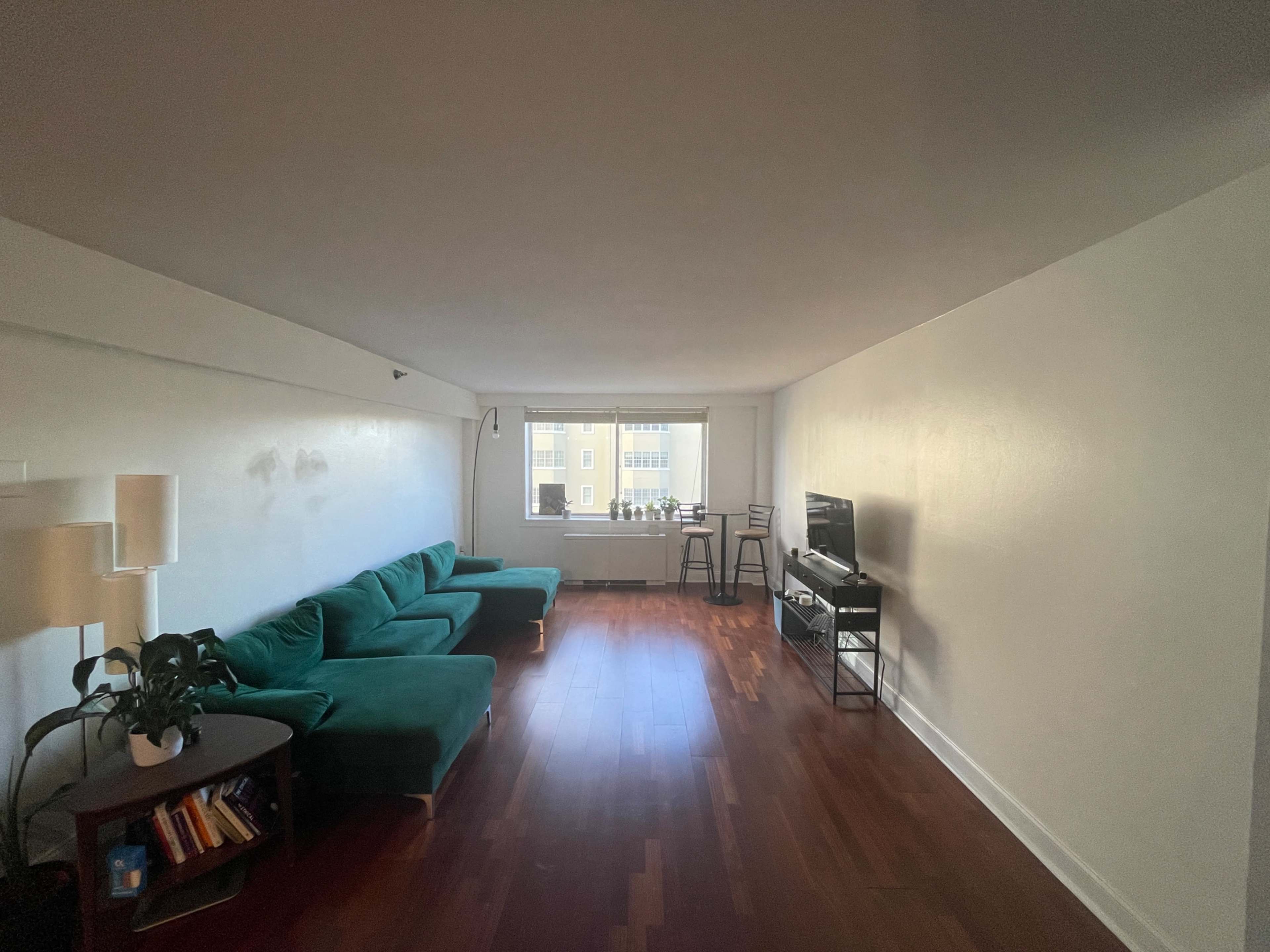 A spacious living room with a green sectional sofa, a small table with books, a TV stand, and a window overlooking a building across the street.