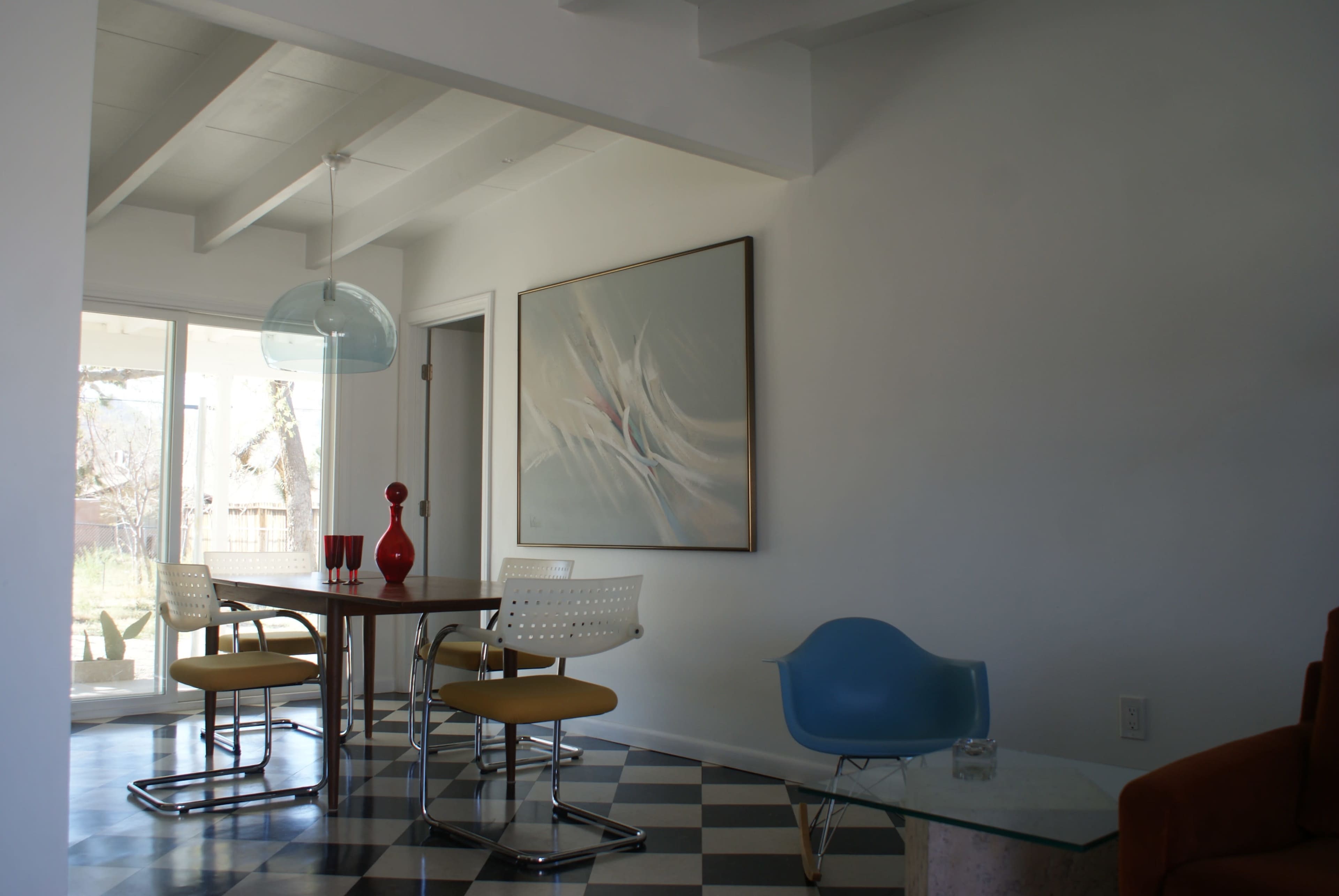A modern dining area featuring a table with chairs, a large abstract painting on the wall, and a blue plastic chair beside a glass coffee table.