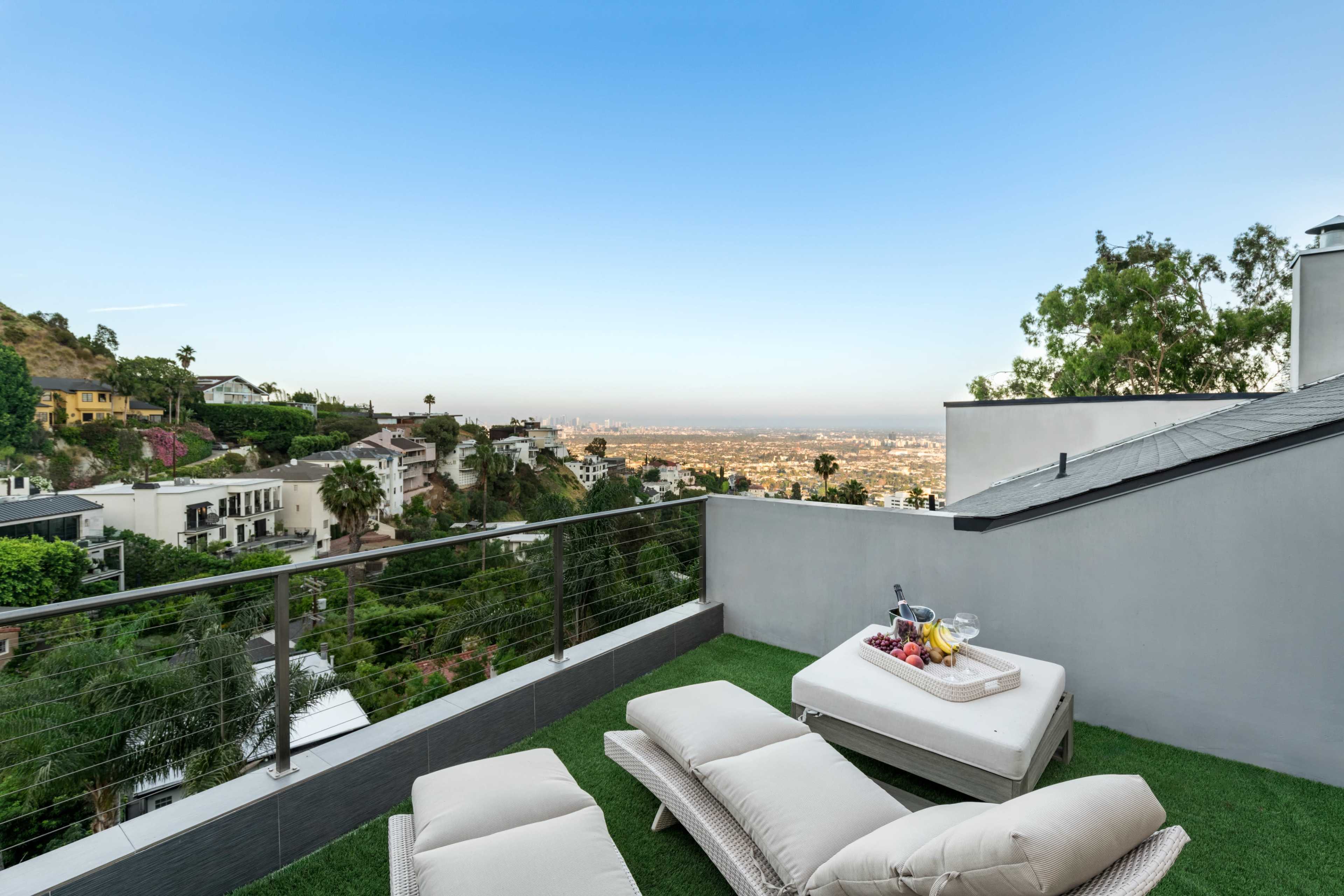 A sunlit terrace features two lounge chairs and a table with fruit, overlooking a cityscape from a hillside.