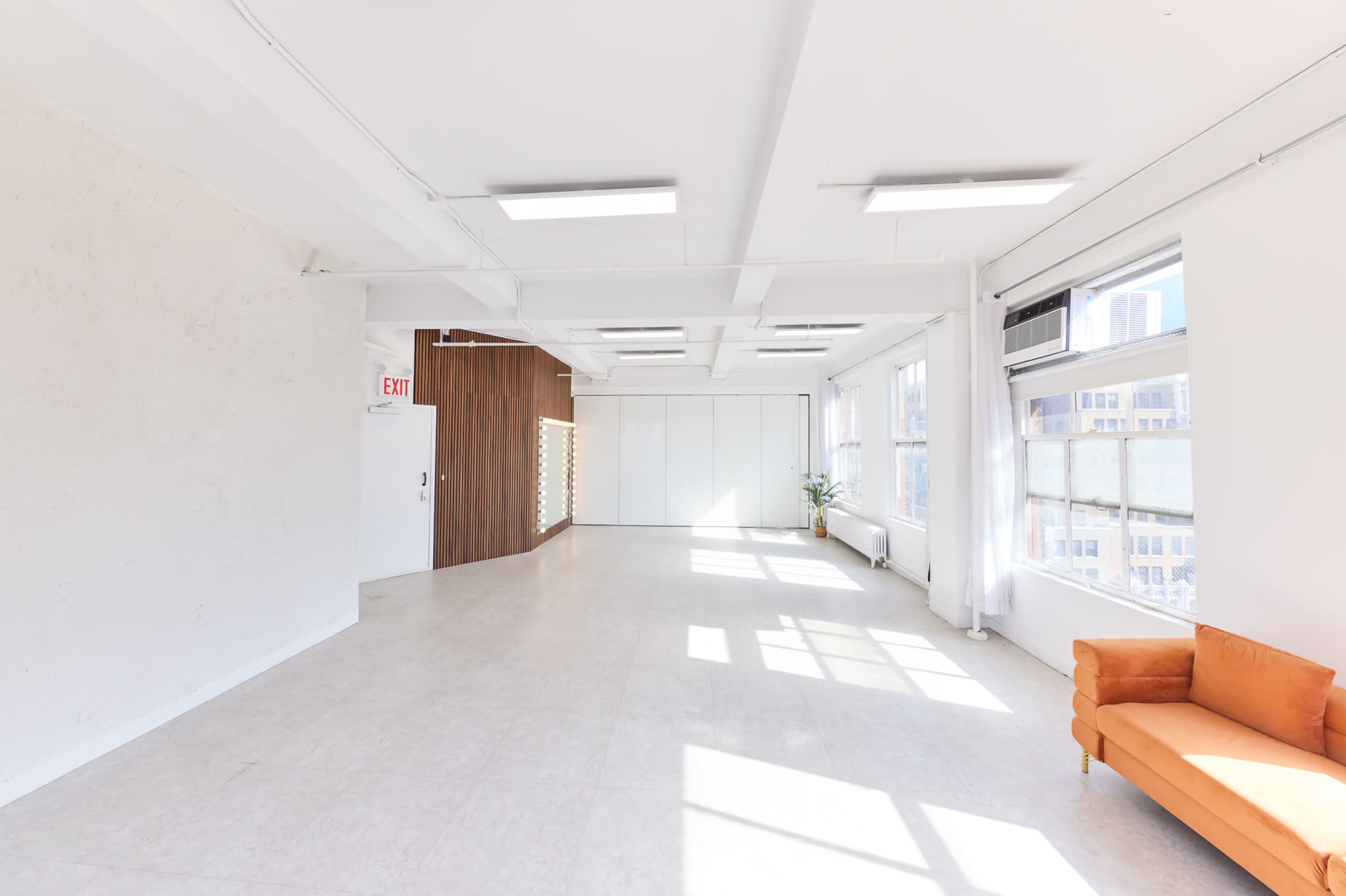The image shows a bright, spacious interior with large windows, light flooring, and an orange couch against one wall.