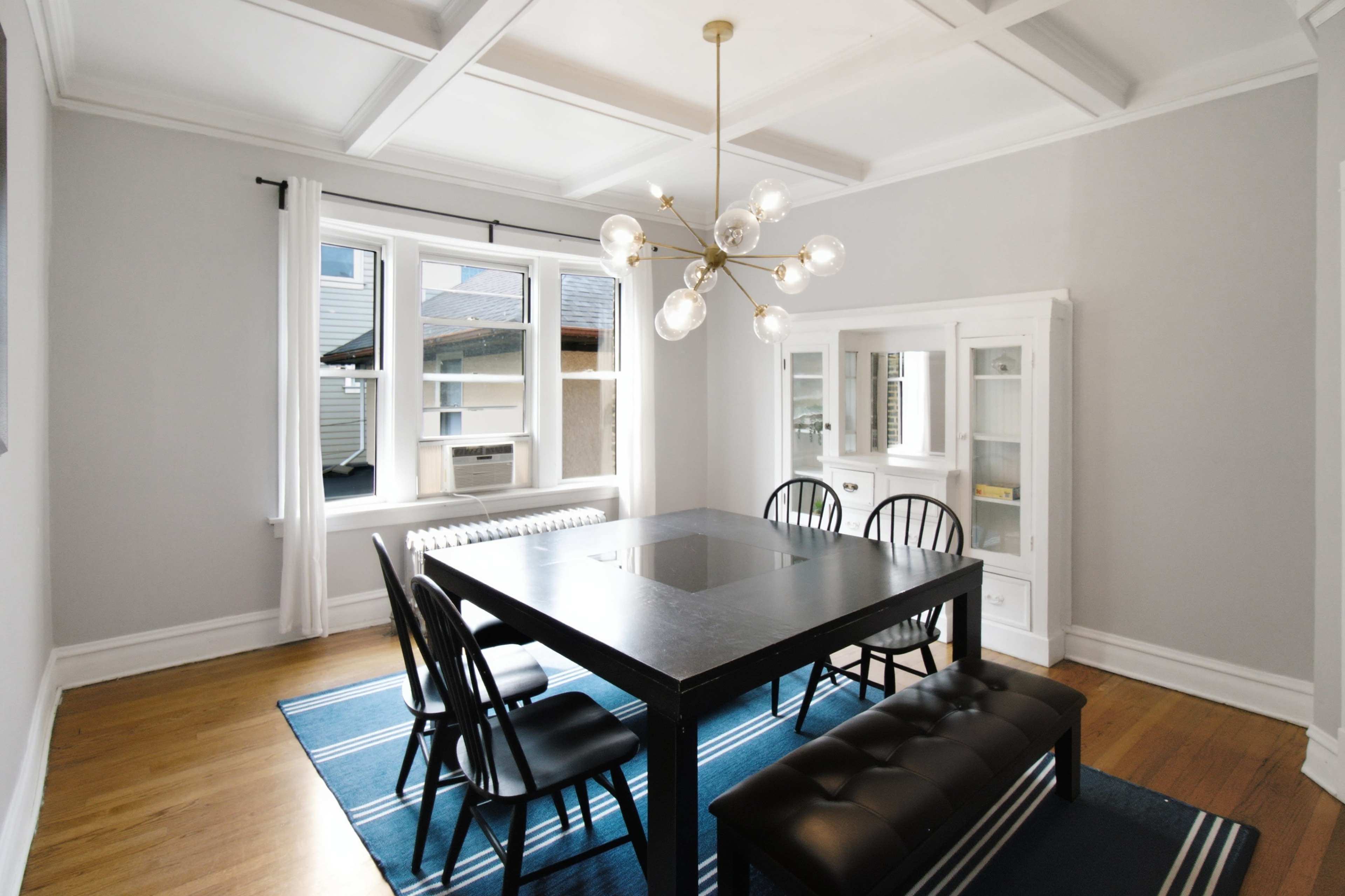 The image shows a modern dining room featuring a black square table surrounded by six chairs on a blue area rug, with a decorative light fixture overhead and a white display cabinet against the wall.