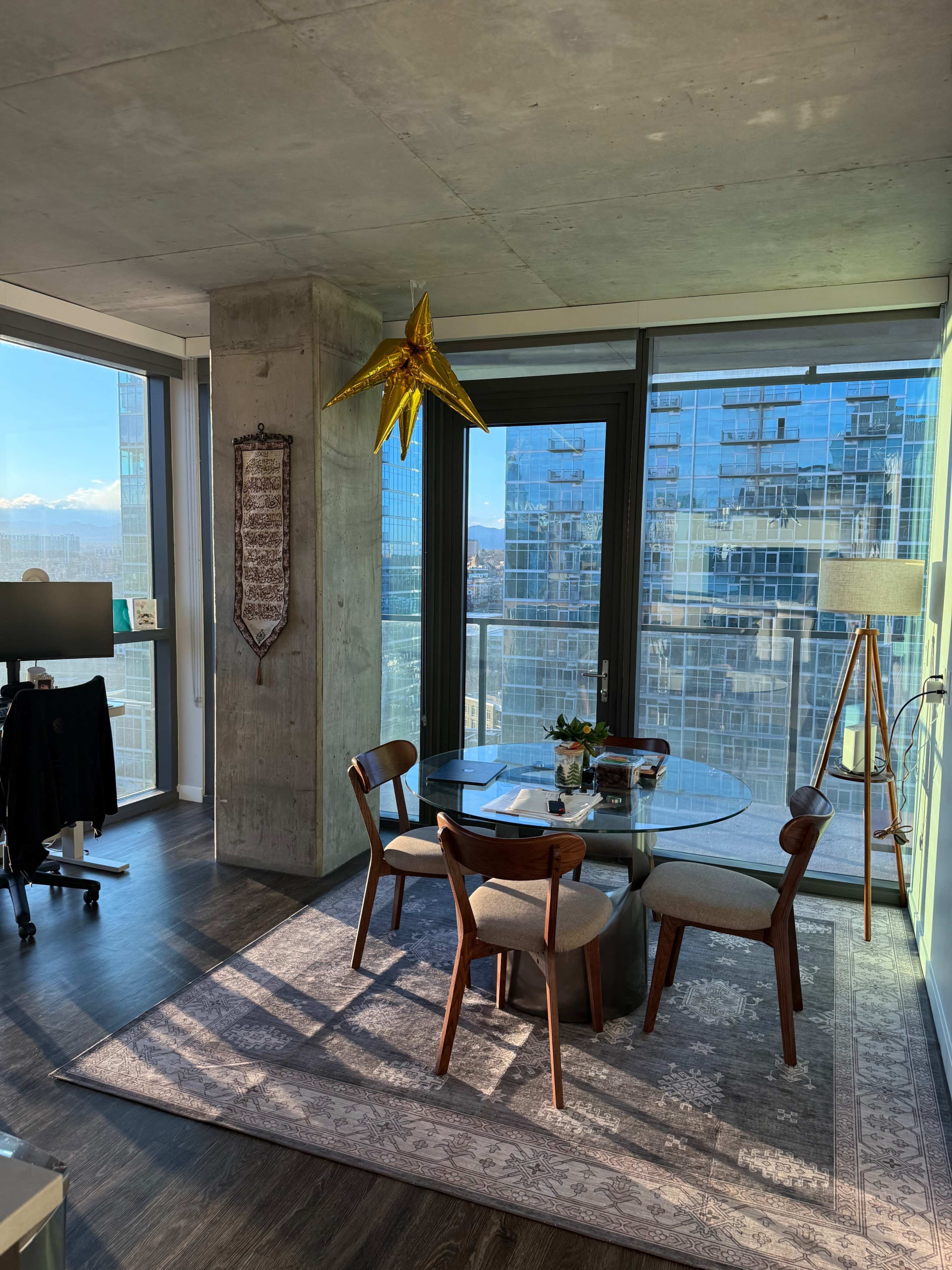 The image shows a modern dining area with a circular glass table, wooden chairs, and a star-shaped decoration, surrounded by large windows showcasing a city view.