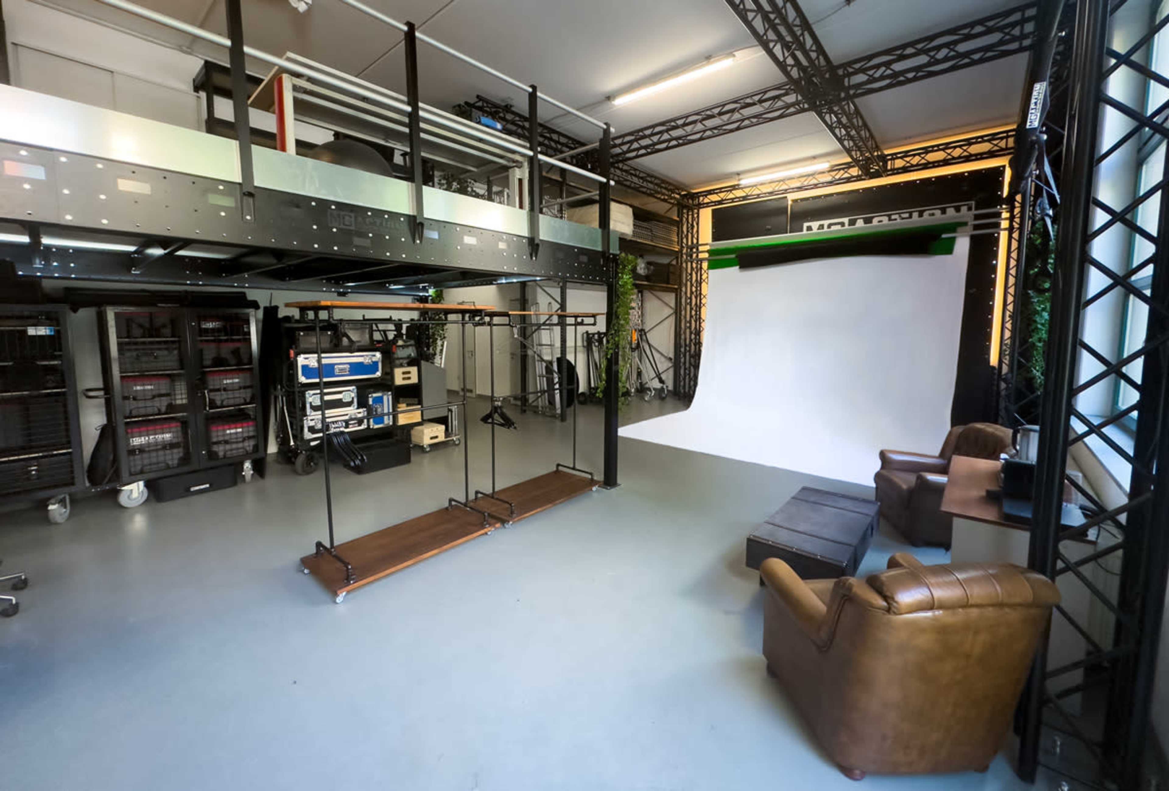 A spacious studio environment featuring a white backdrop, a seating area with brown chairs, and various technical equipment organized on shelves.