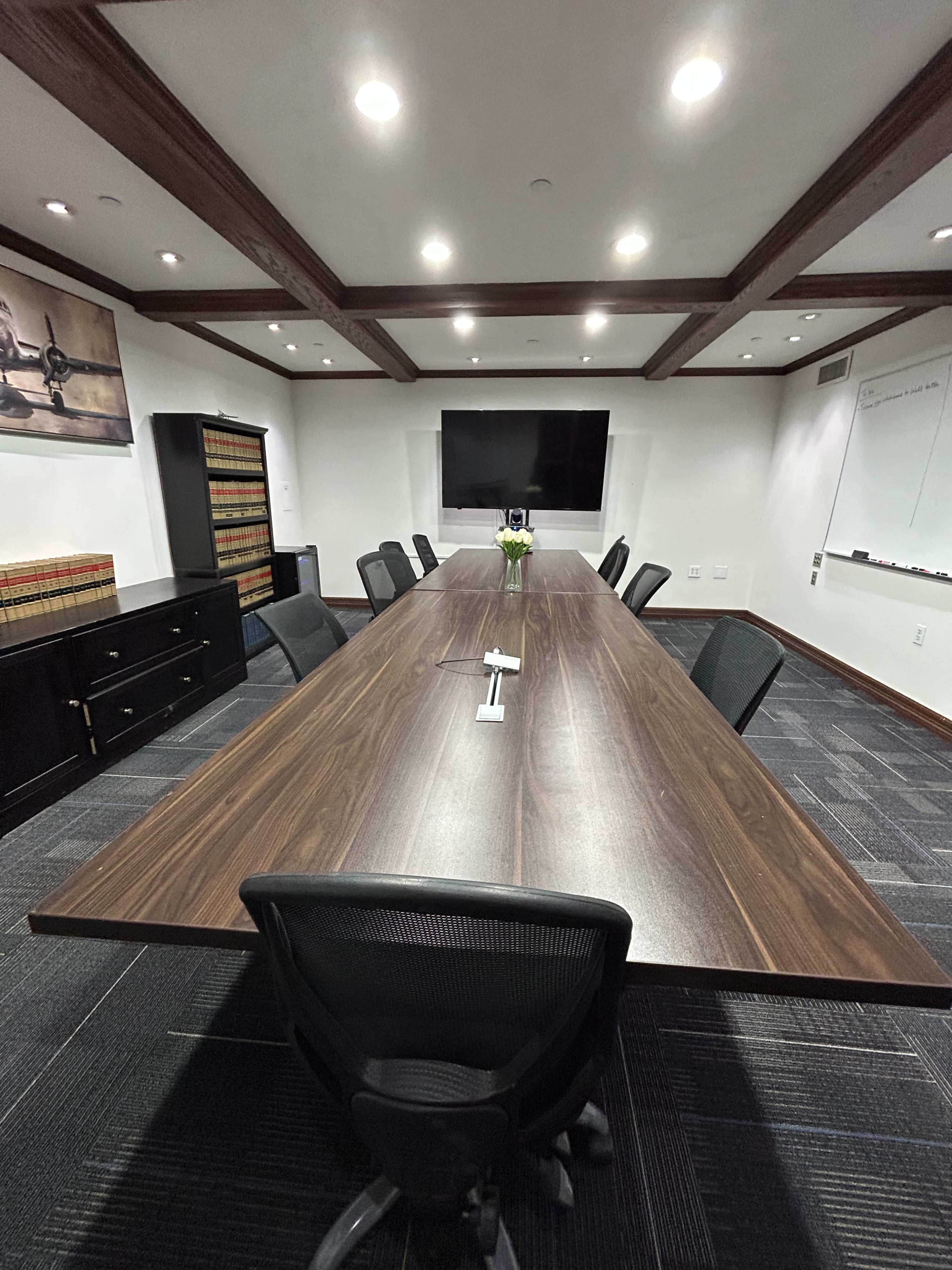A large conference room features a long wooden table surrounded by black office chairs, with a wall-mounted screen and shelves containing legal books in the background.